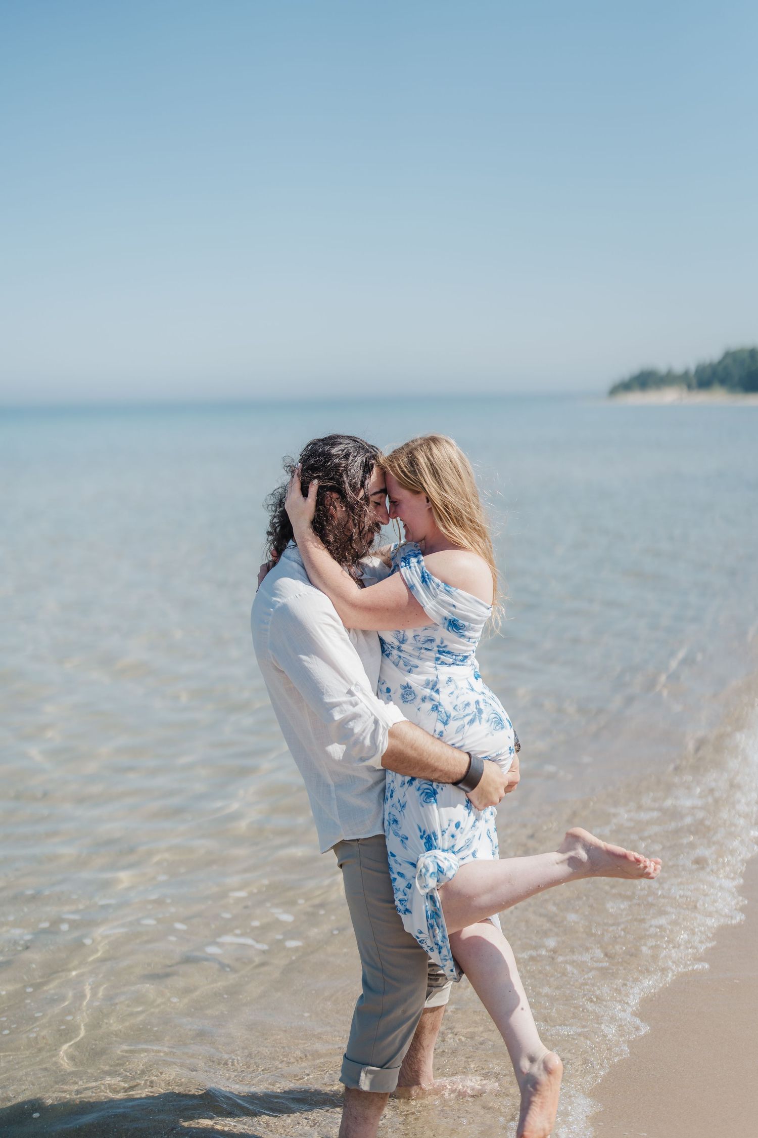 A romantic couple embraces while standing in shallow ocean water on a sunny beach.