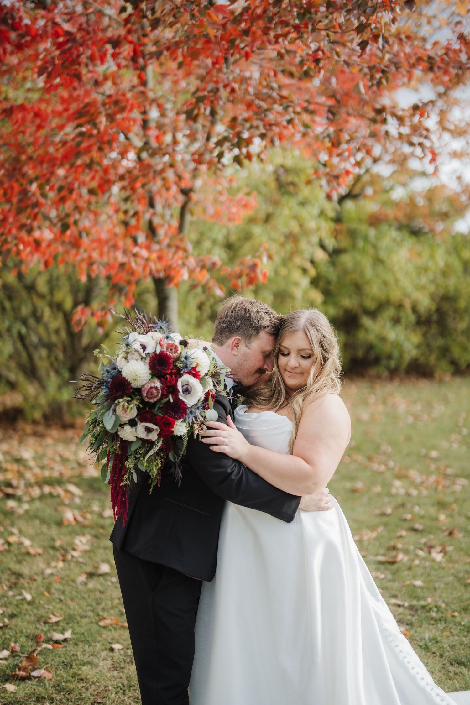 A couple embraces under vibrant red autumn foliage while holding a burgundy and white wedding bouquet.