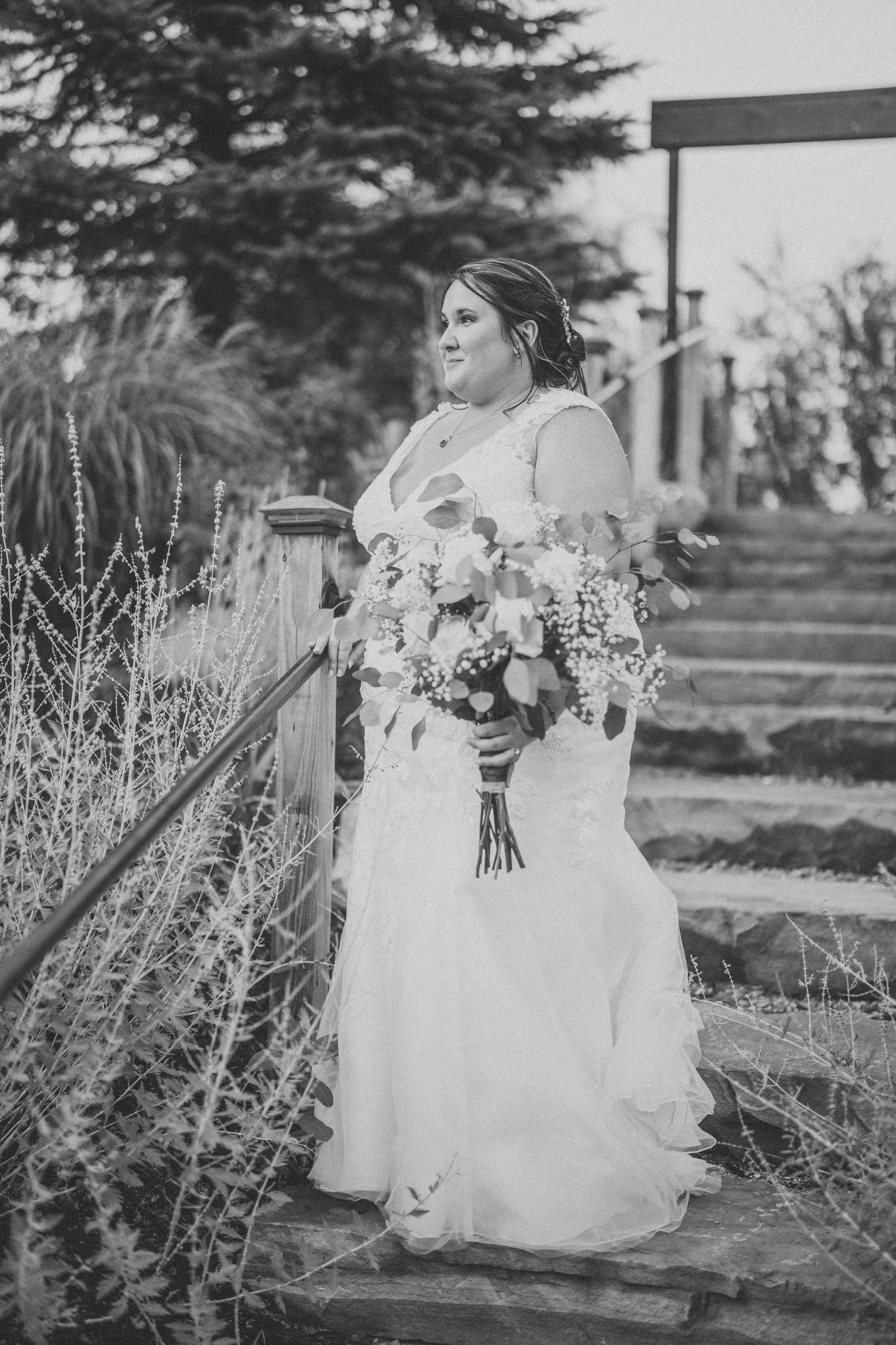 A bride in a white wedding gown stands on stone steps holding a beautiful floral bouquet in black and white.