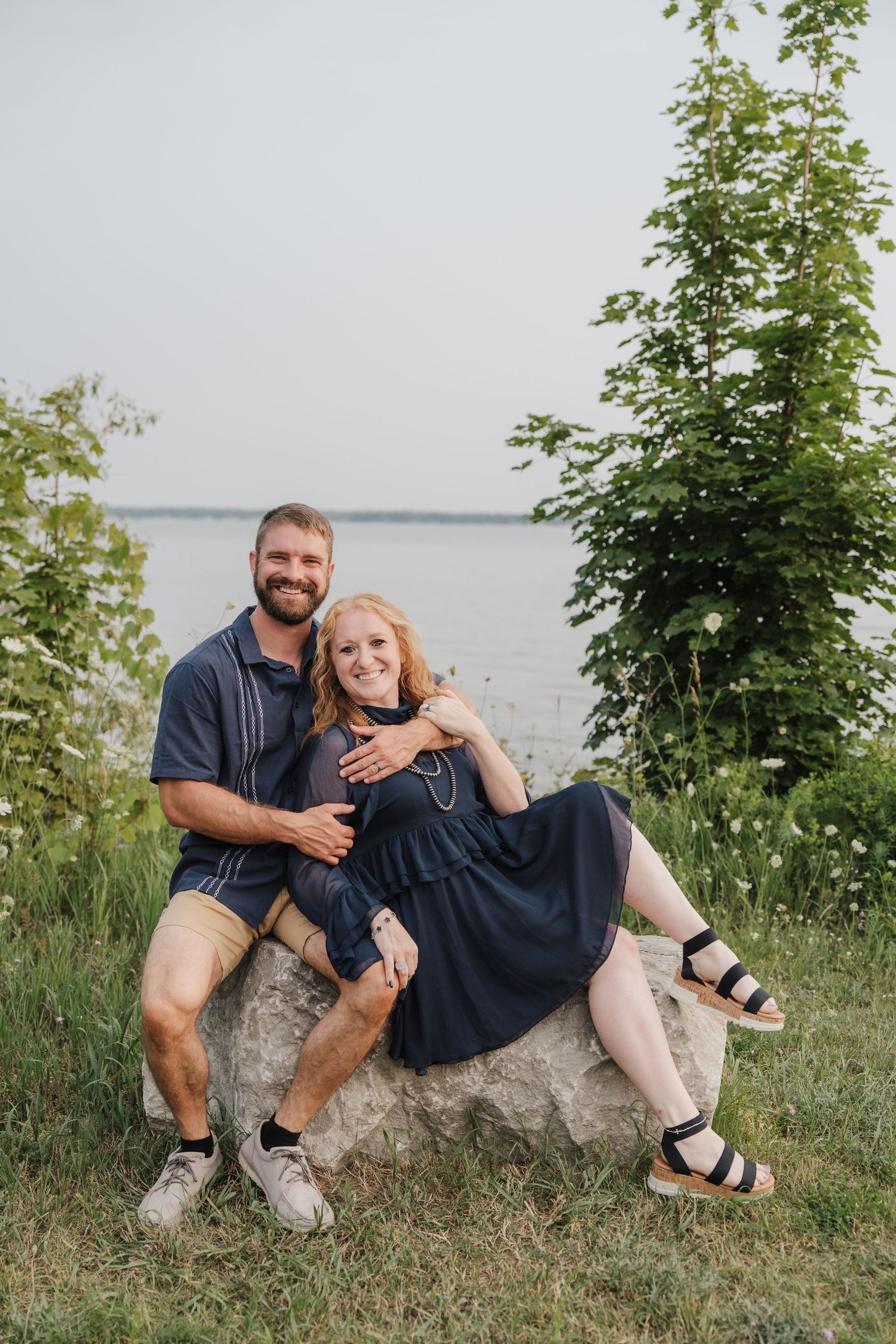 Smiling couple sitting on a large rock near a lakeside, man in navy shirt and shorts, woman in navy dress and sandals.