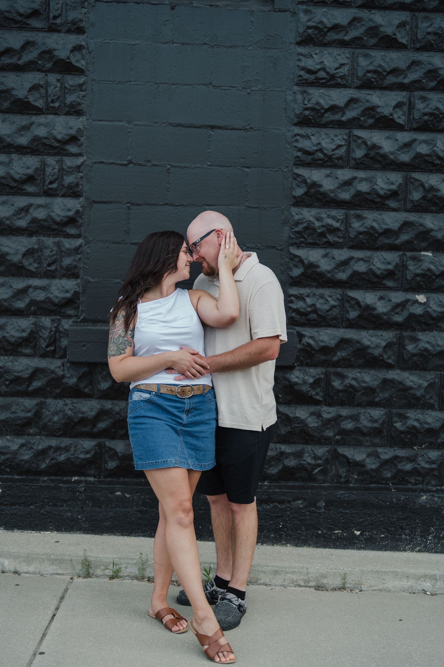 Couple sharing a romantic moment against a black brick wall outdoors, woman in white top and denim skirt, man in beige shirt.
