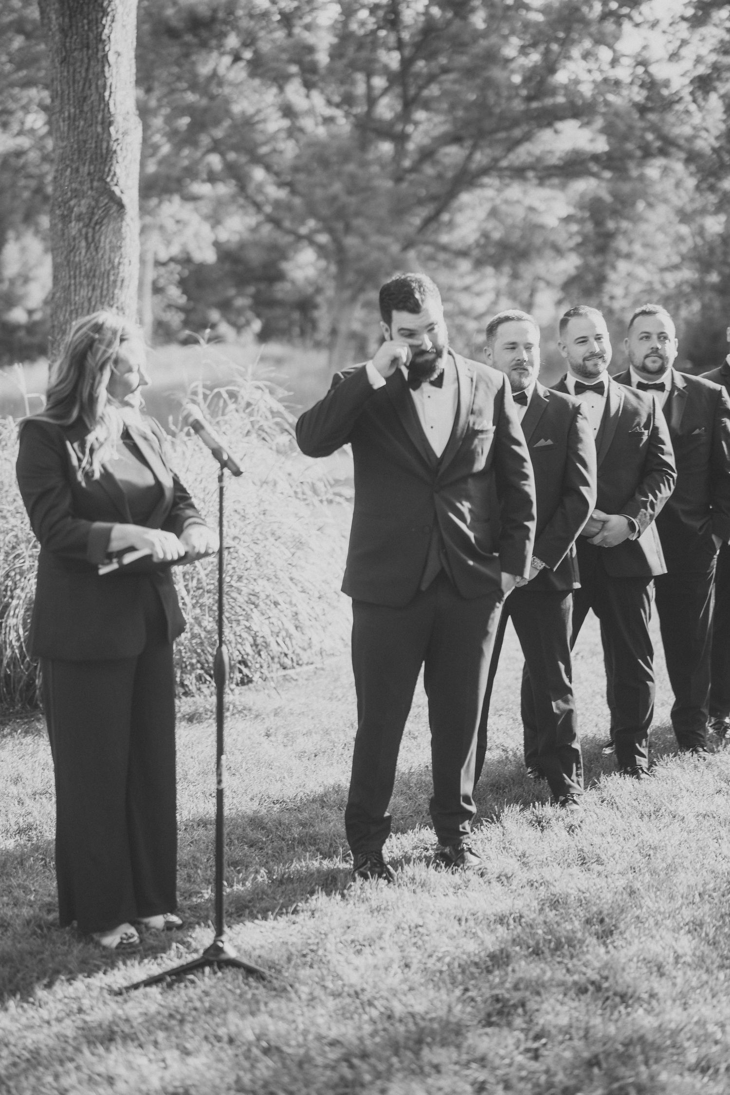 Wedding party in black attire poses on grassy hillside under tall trees.