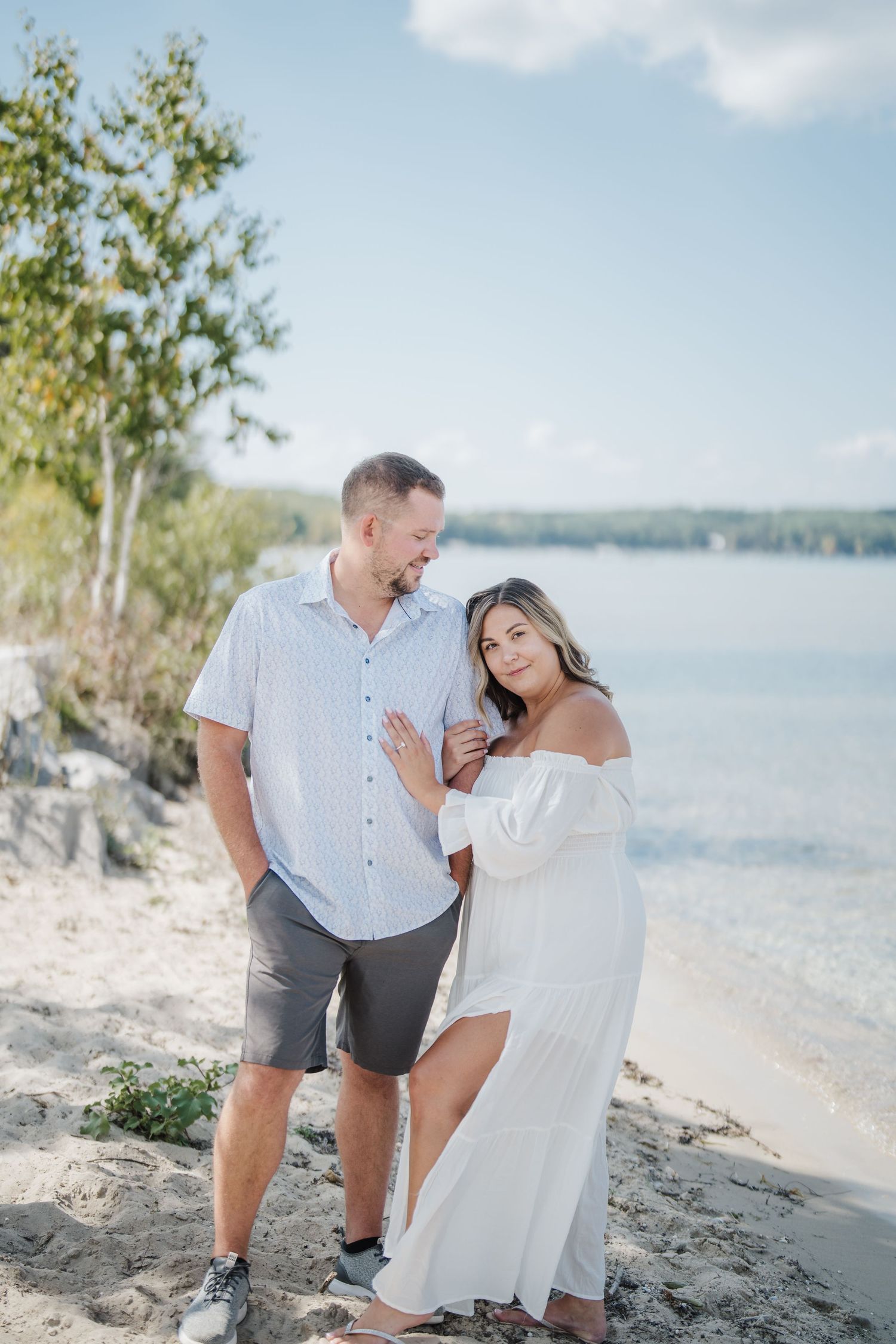 A couple shares romantic moments together on a scenic beach with water and trees in the background.