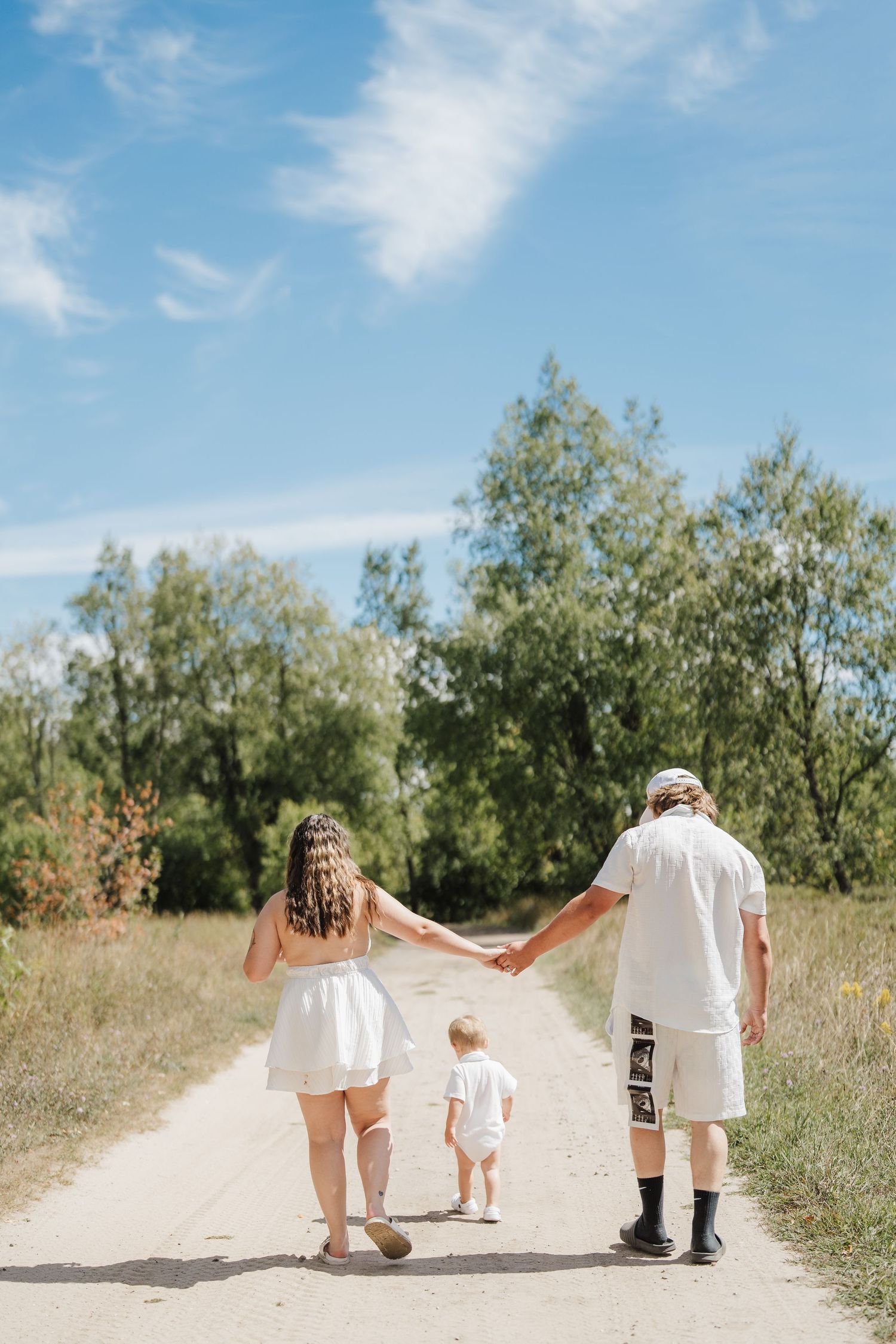 A family walks hand in hand down a sunny dirt path bordered by green trees and bushes on a summer day.