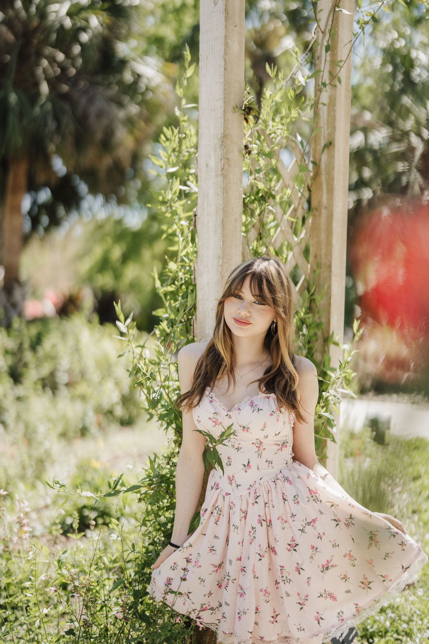 A floral sundress twirls gracefully in a sunlit garden setting among green foliage and trees.
