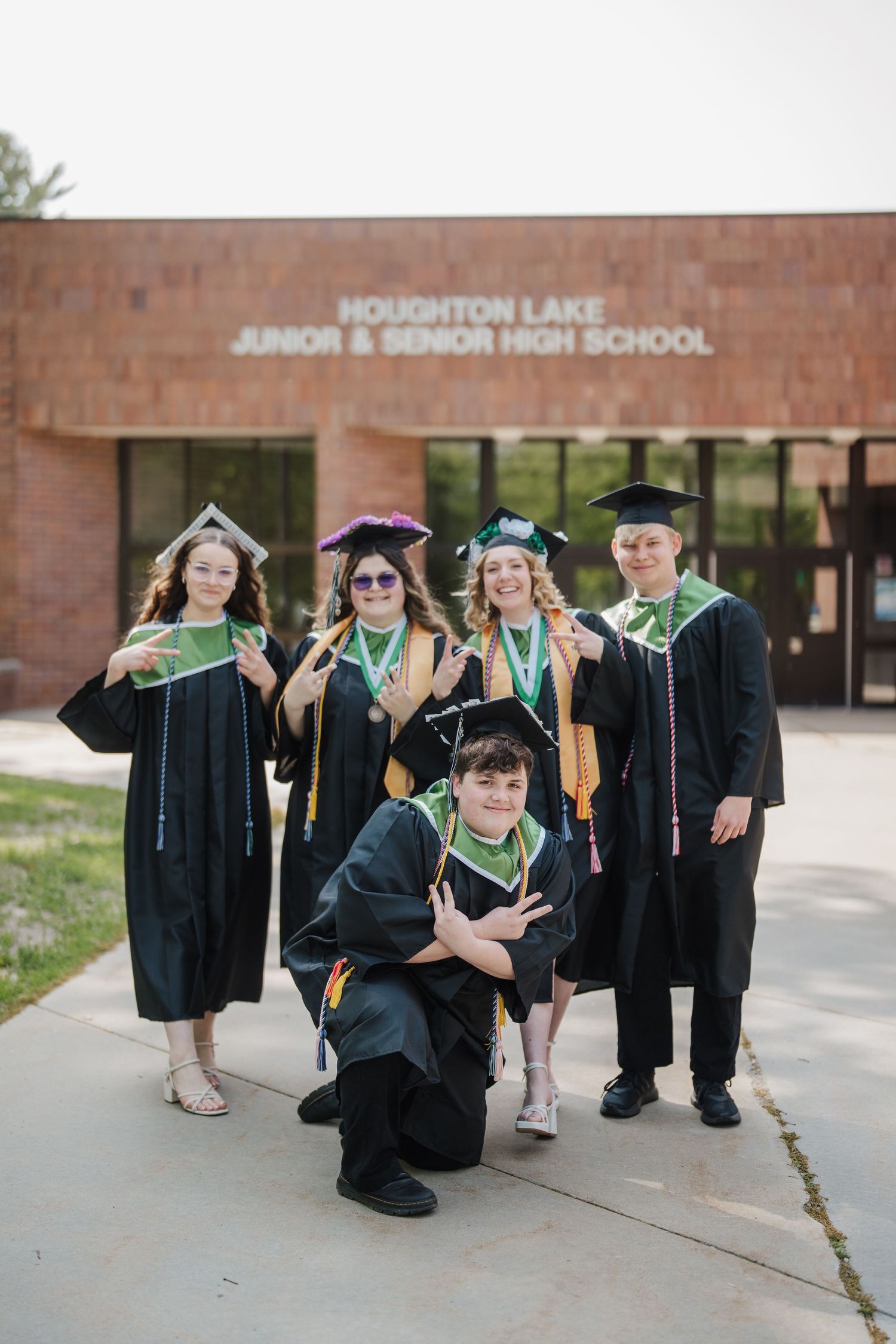 Graduates in black caps and gowns pose in front of Houghton Lake Junior & Senior High School building.