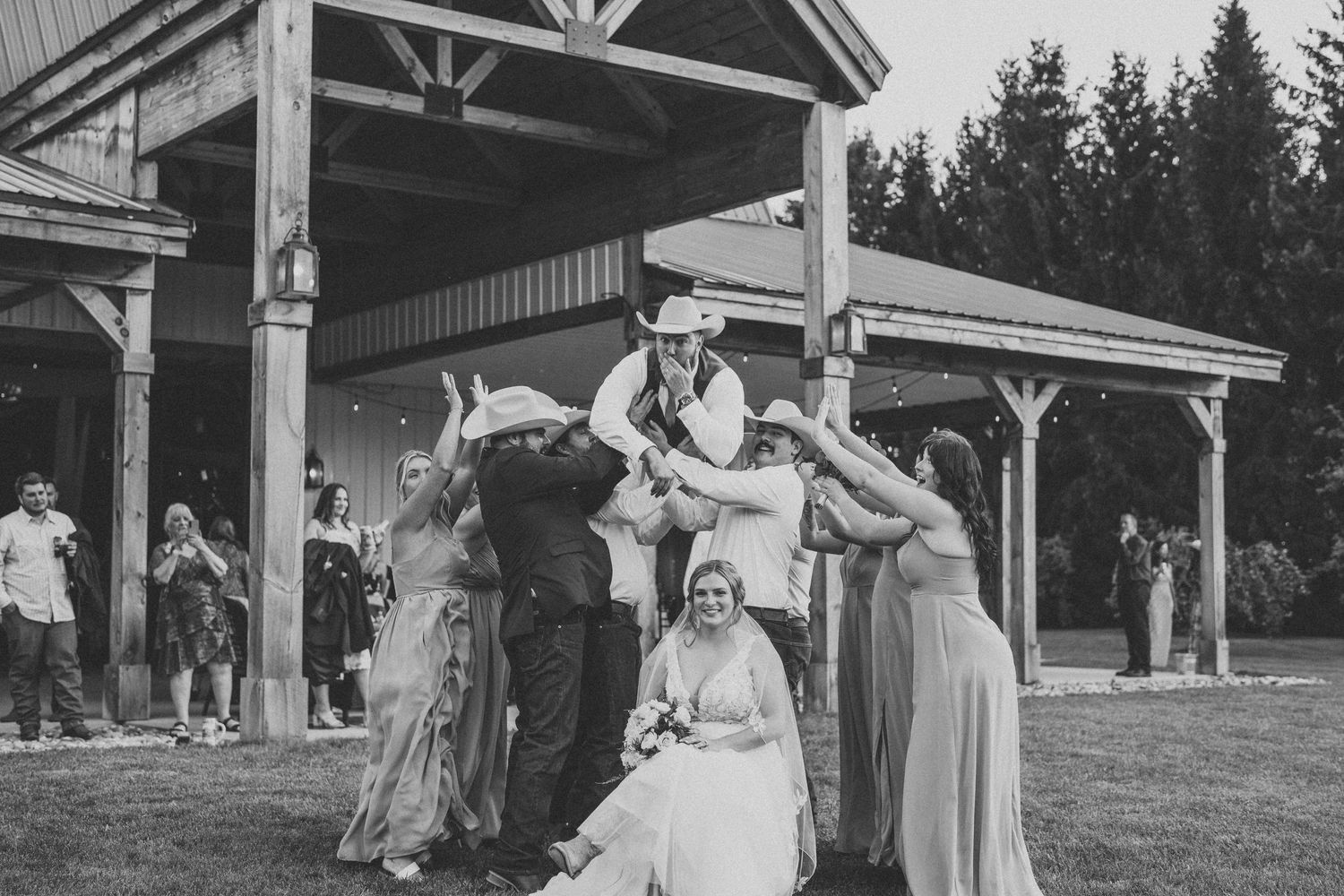Wedding party poses playfully under wooden pavilion in black and white photography.