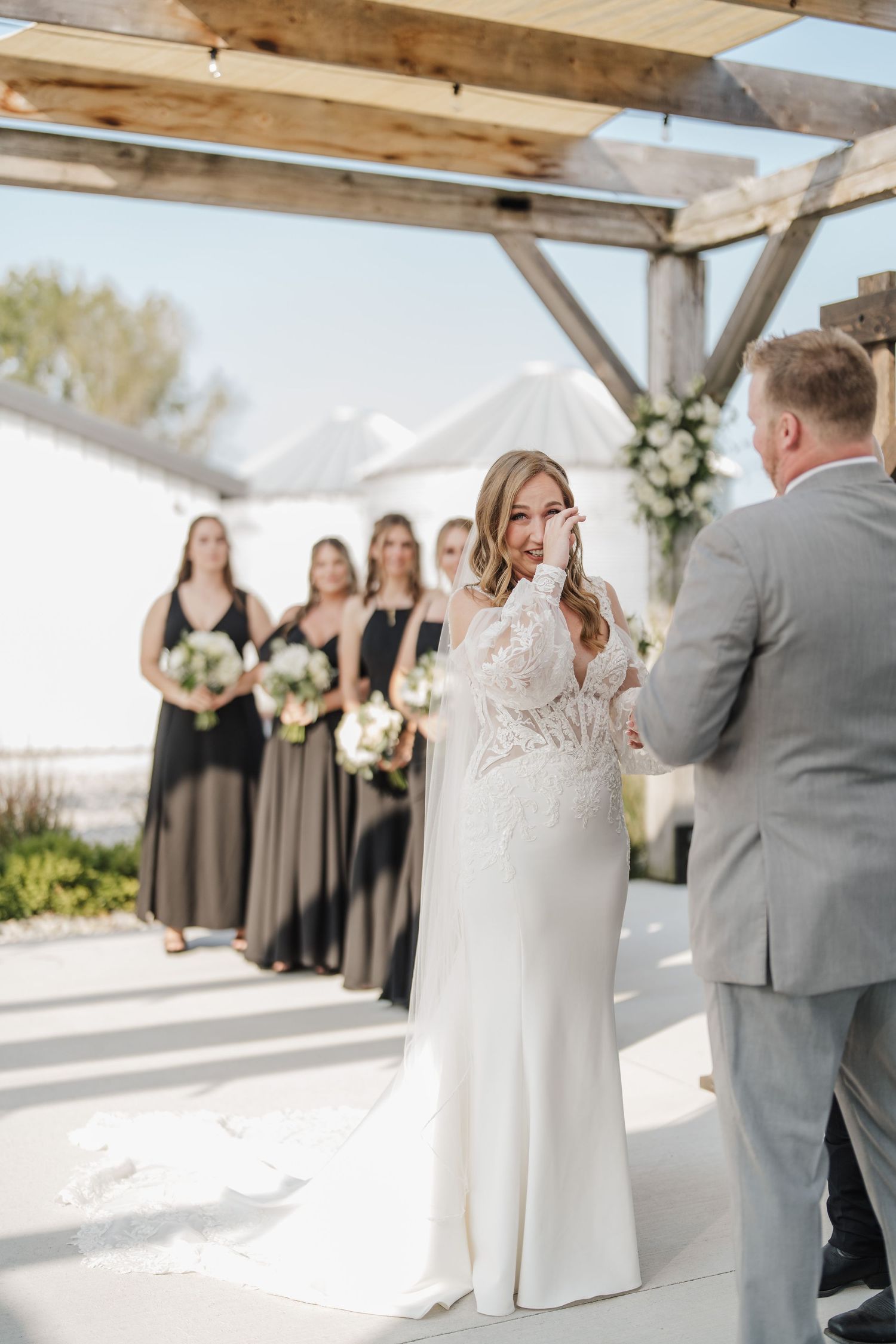 A wedding ceremony under a wooden pergola with bridesmaids in black dresses.
