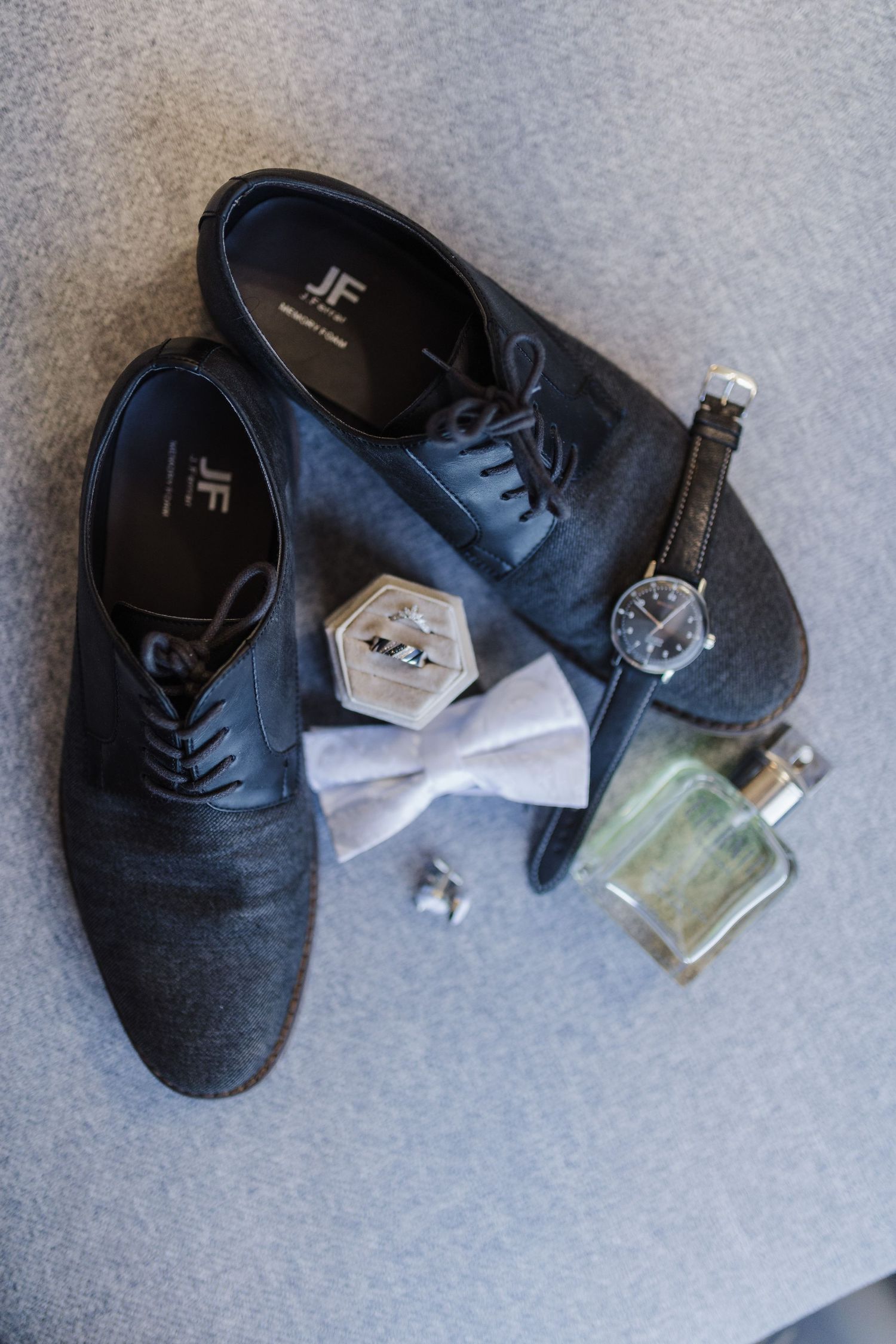 Groom's accessories including black dress shoes, bow tie, watch and cologne on gray surface.