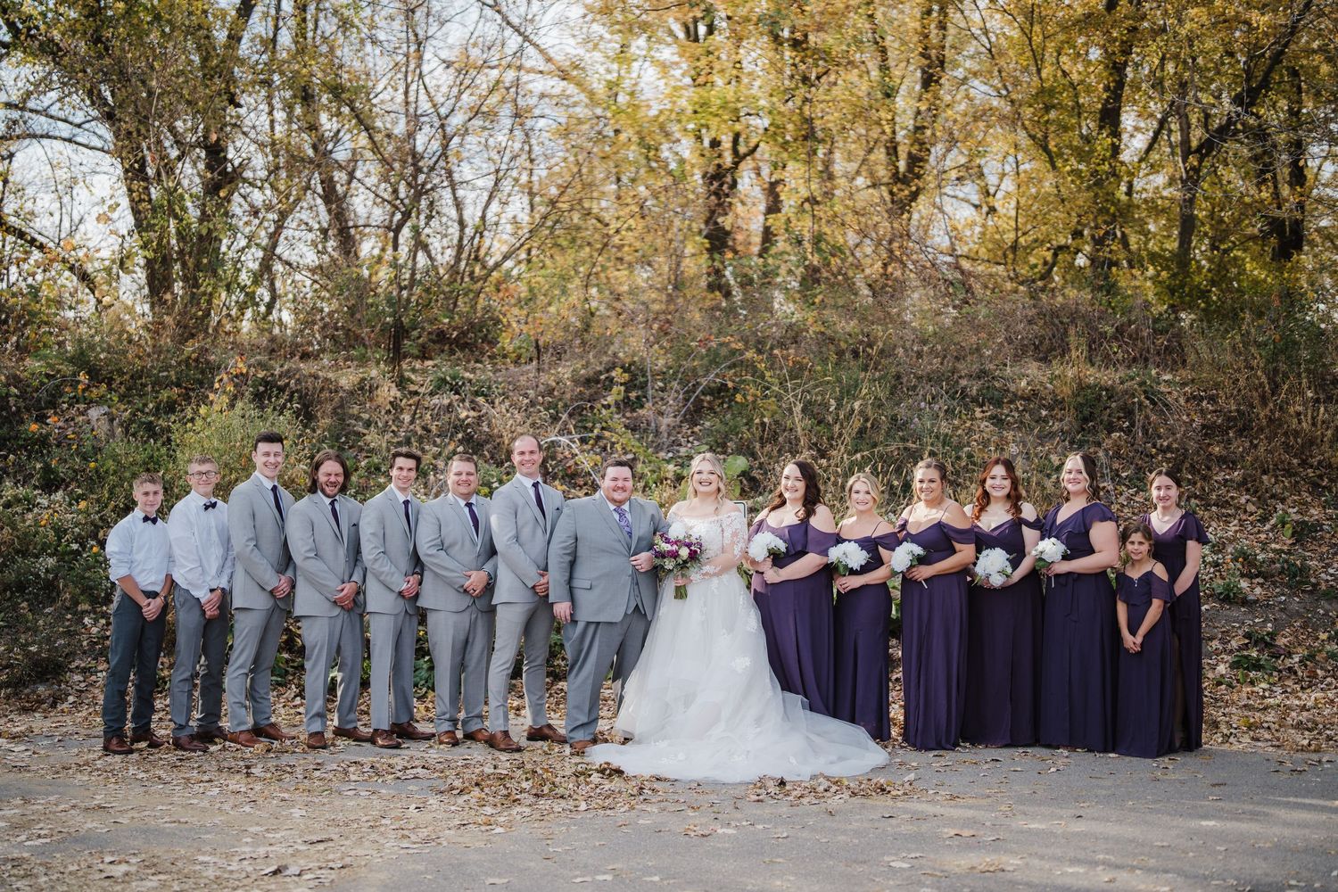 Wedding party in gray suits and purple dresses poses outdoors during fall season.