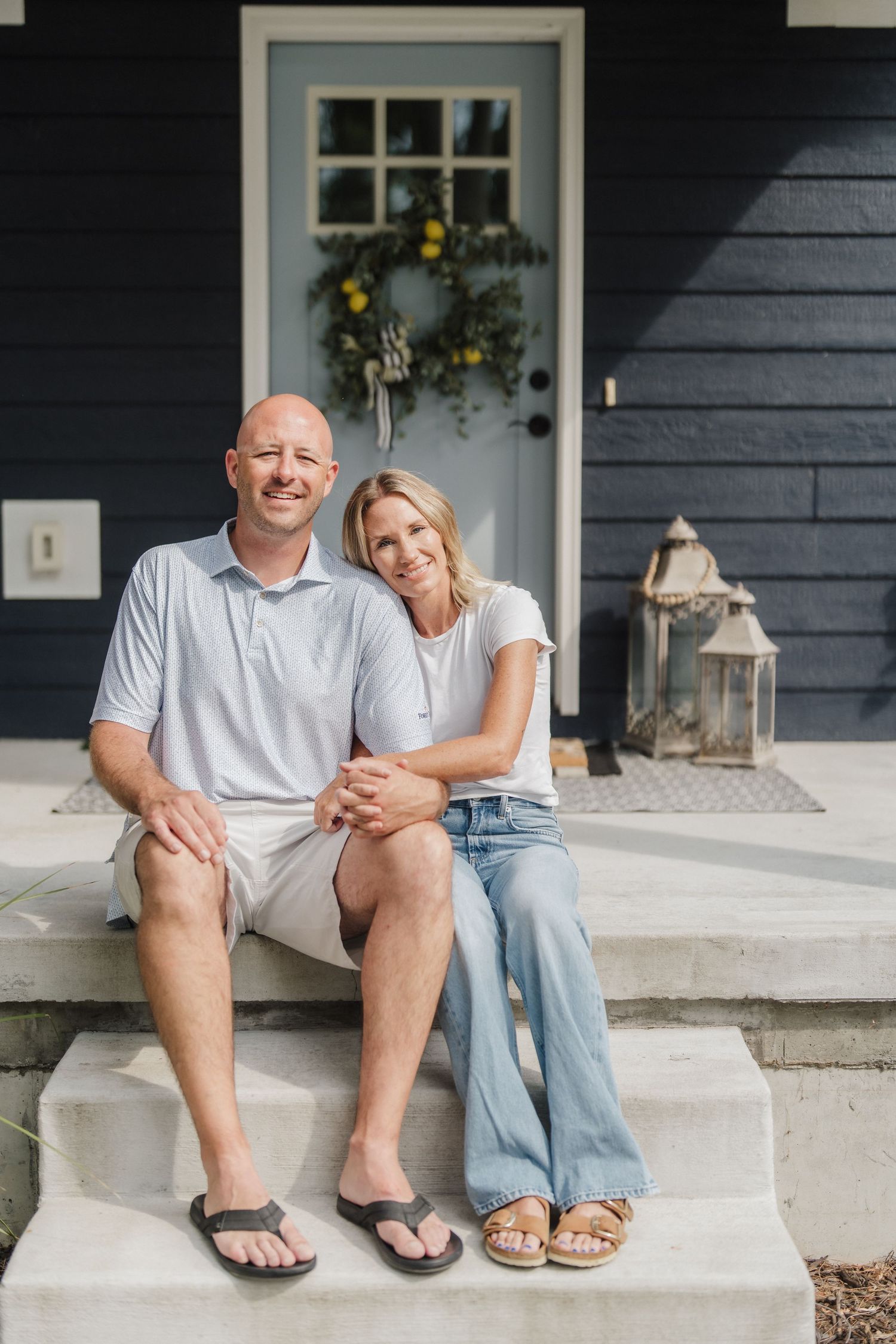 A smiling couple sits together on front porch steps of a navy blue home with a wreath on the door.