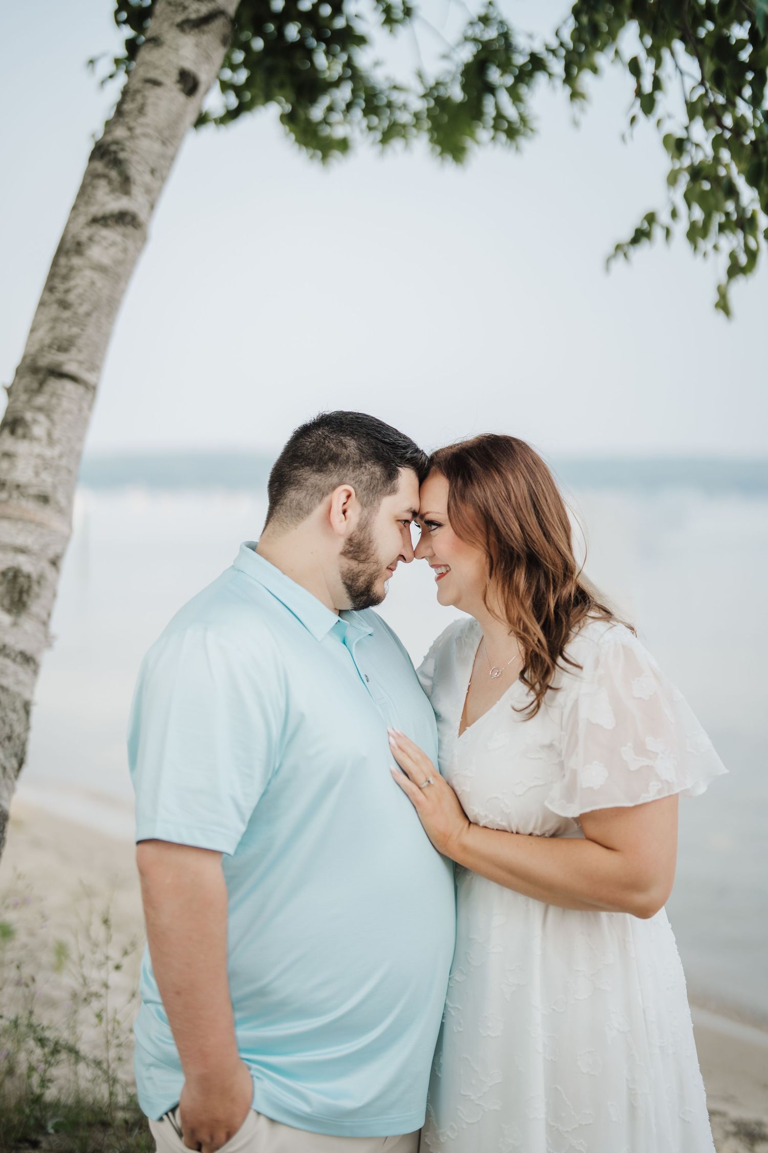 A romantic couple shares tender moments near birch trees along a lakeside beach in soft natural lighting.