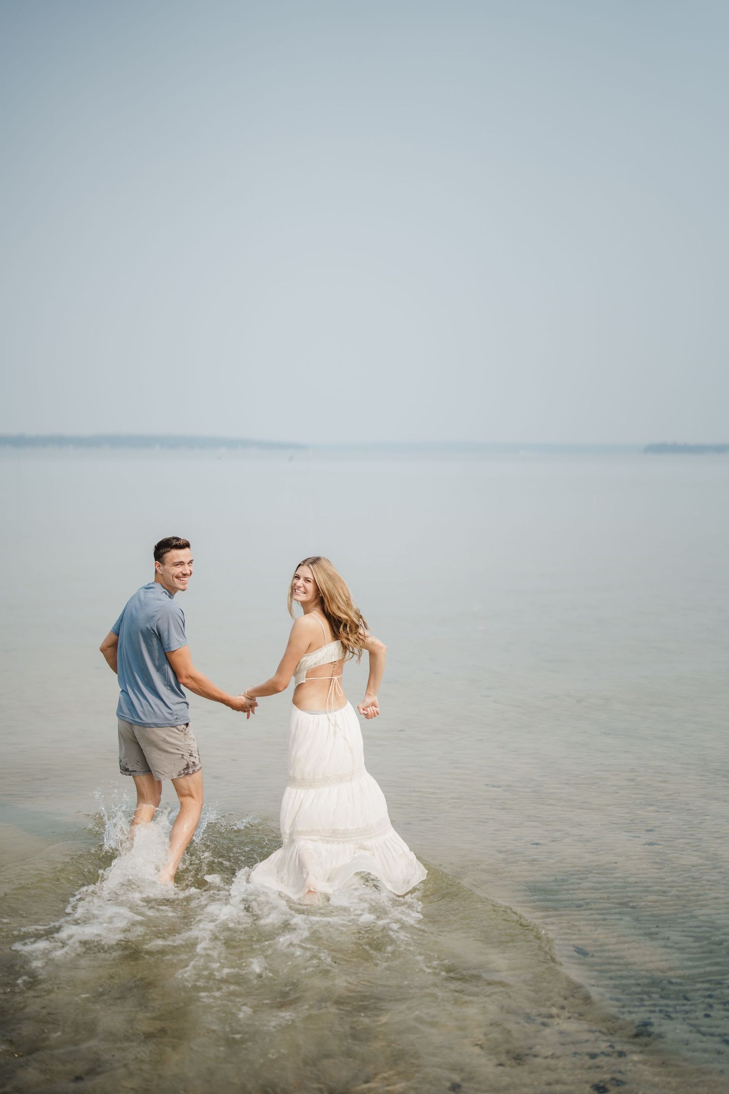 A couple splashes through shallow coastal waters on a misty day, with the person in a flowing white dress.