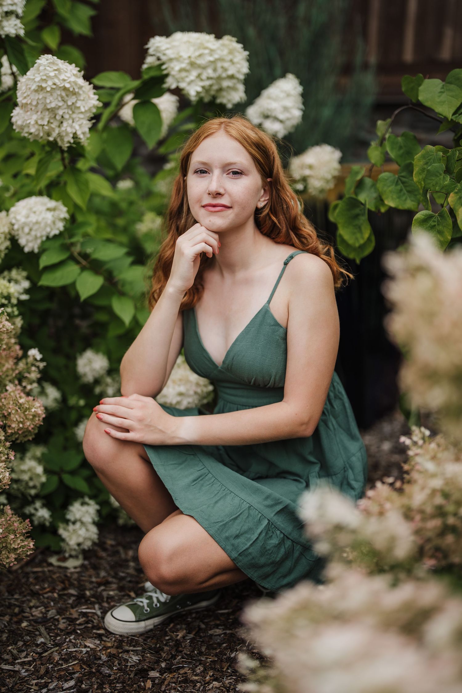 A person in a green dress poses thoughtfully among blooming white hydrangea flowers in a garden setting.