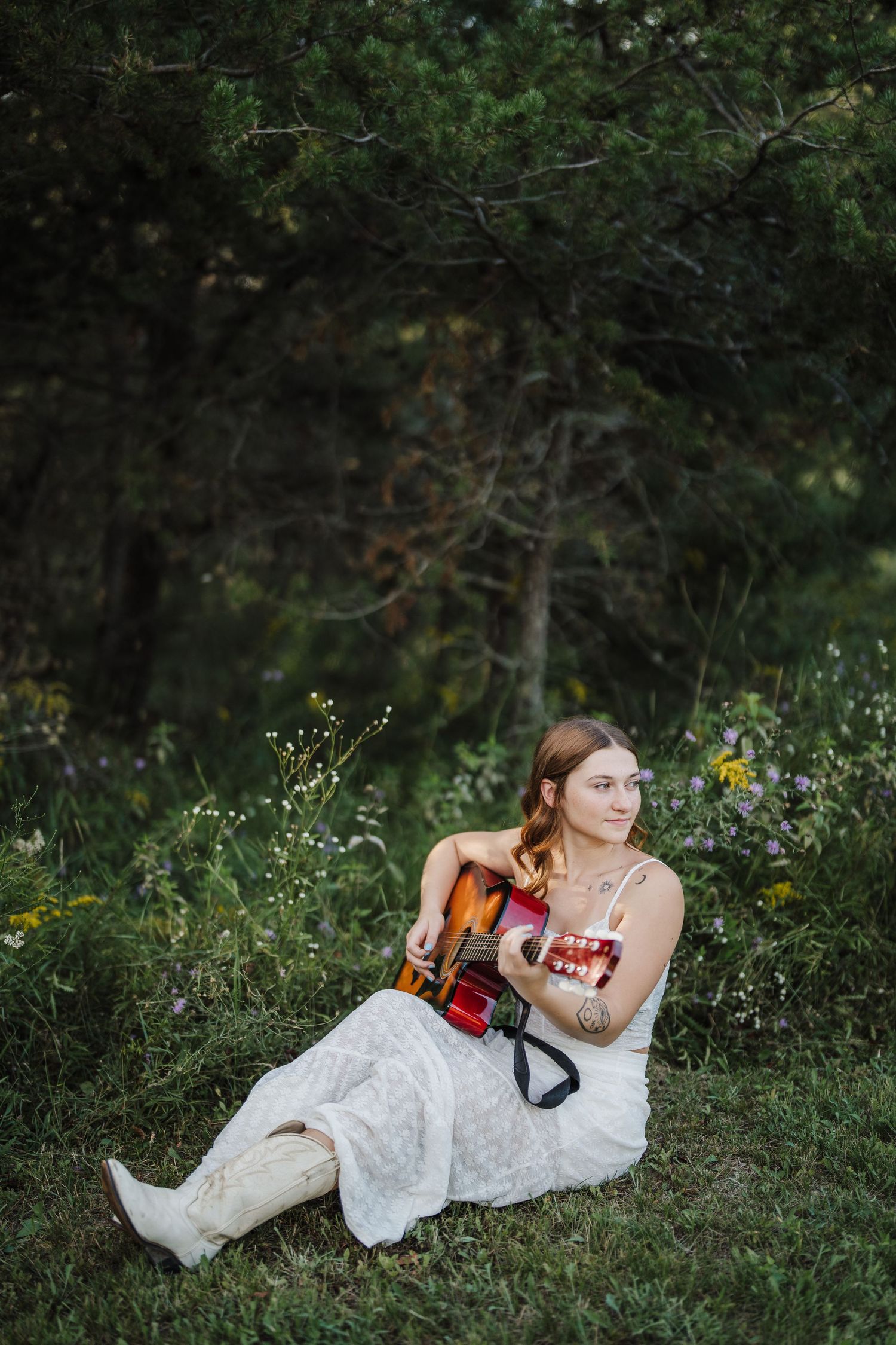 A person in a white dress and boots sits on grass with a violin surrounded by wildflowers and trees.