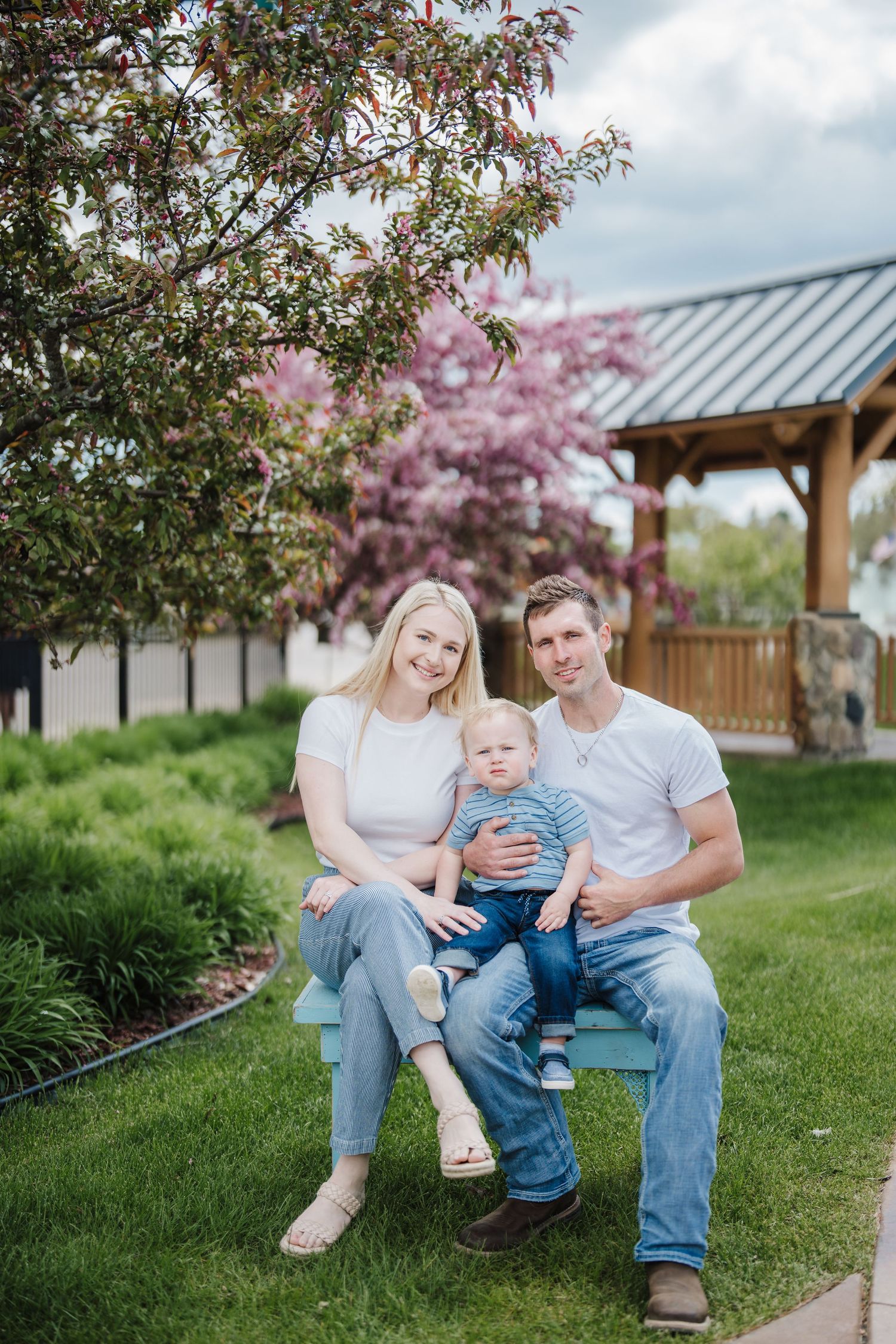 A family sits together on a grassy hill near a gazebo with blooming cherry trees in the background.