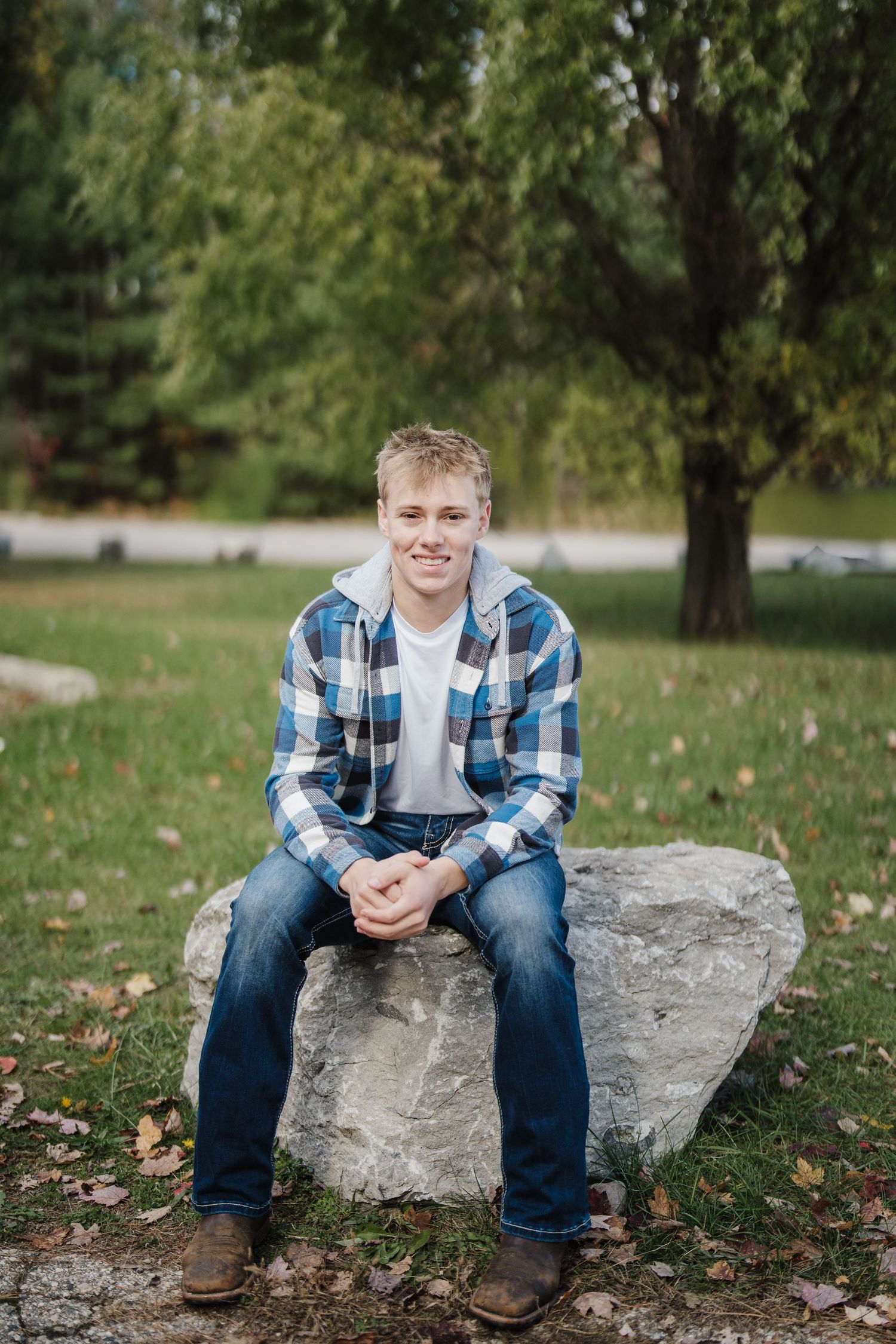 Casual outdoor portraits of someone sitting on a large rock in a park wearing a plaid shirt and jeans.