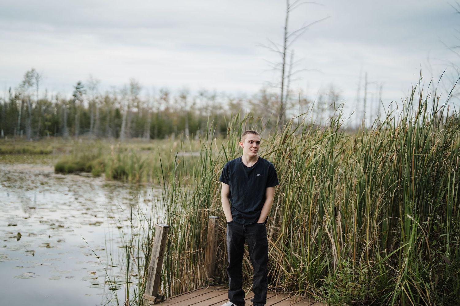 Person in black shirt stands among tall reeds and marsh grasses near calm water at sunset.