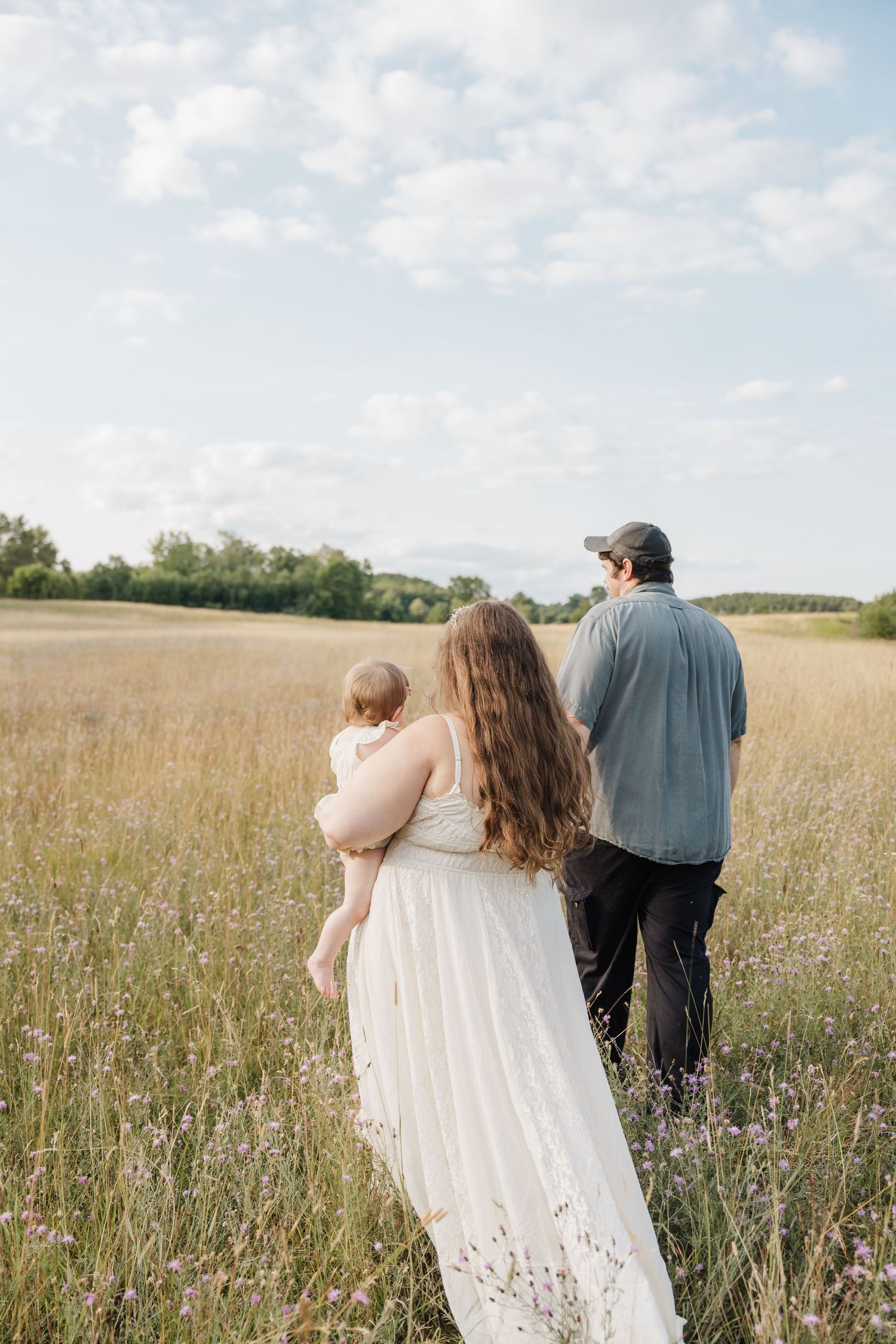 Family of three walking through a wildflower meadow, mother in white dress holding baby, father in gray shirt and cap.