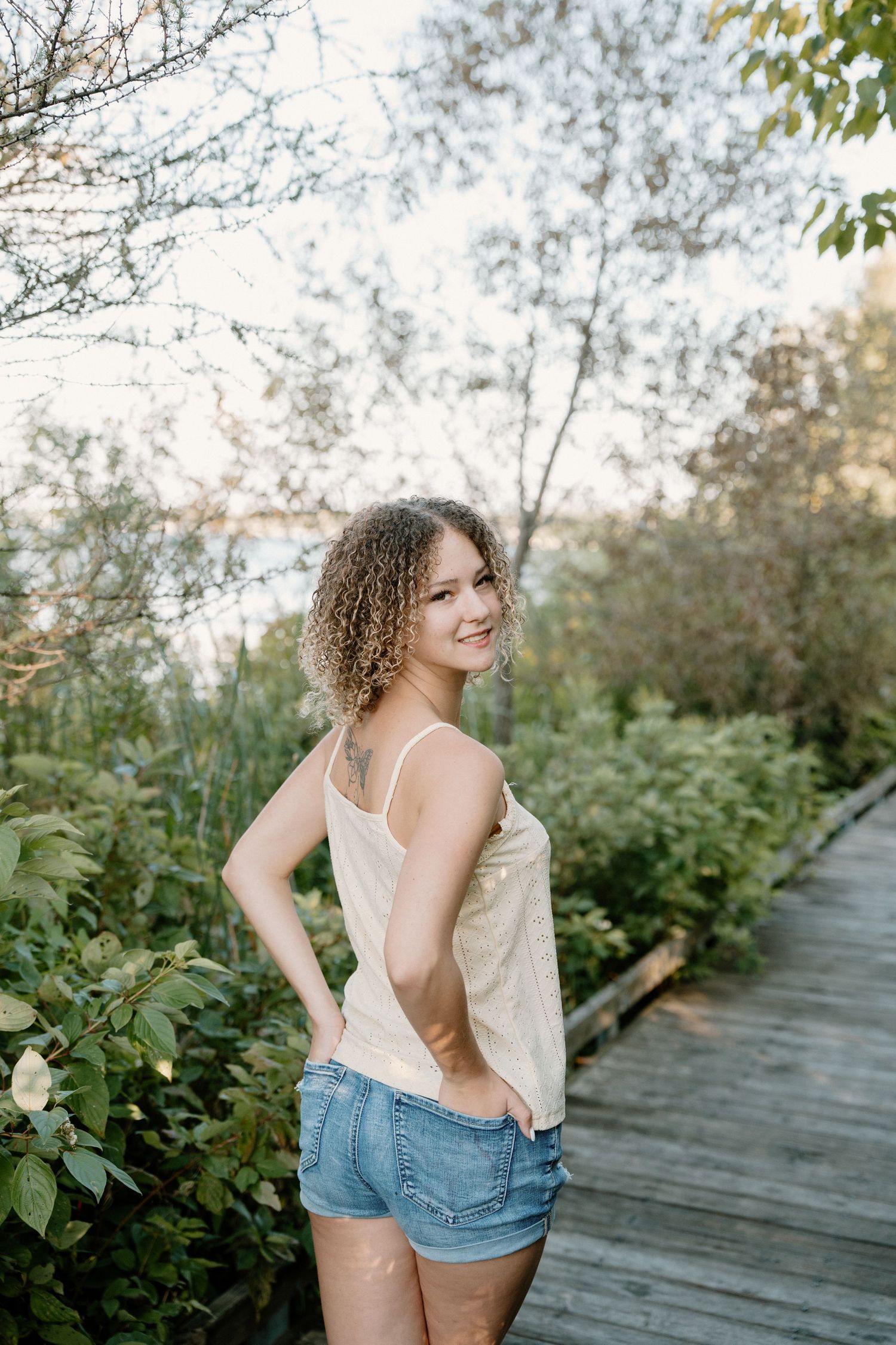 Young woman with curly hair in denim shorts and tank top smiling on a wooden boardwalk surrounded by greenery.