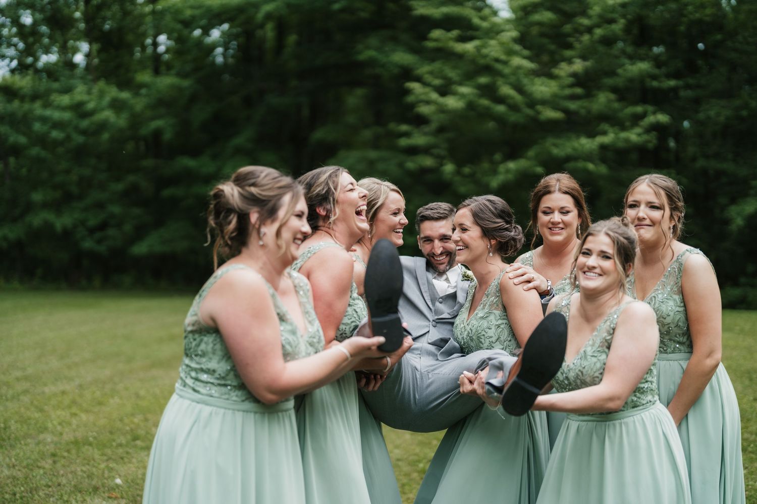 Bridesmaids in mint dresses share laughter while carrying groomsman outdoors.