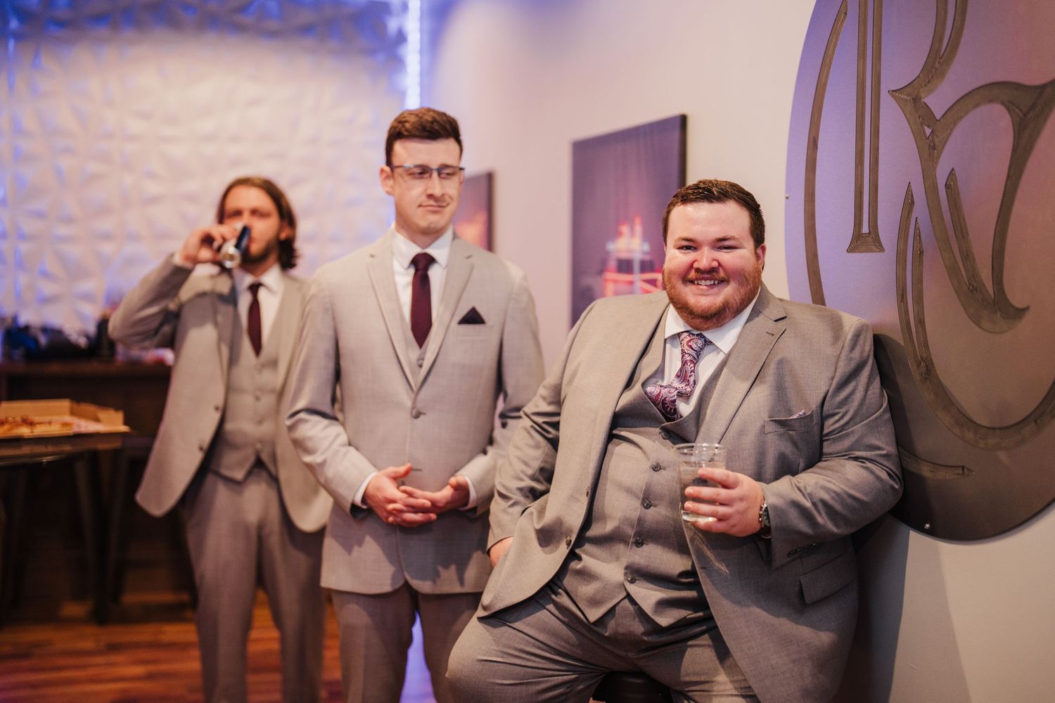 Groomsmen in light gray suits gather for candid moments at an indoor wedding venue.