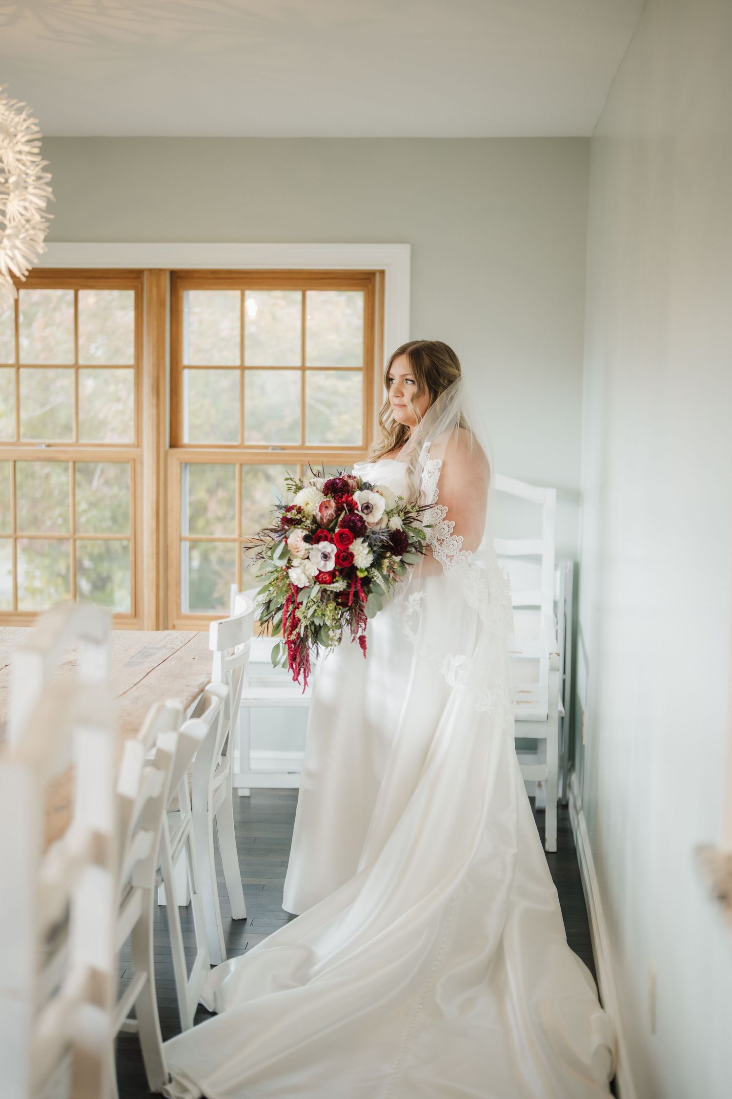 A bride in a flowing white gown holds a stunning burgundy and blush bouquet while standing by a sunlit window.