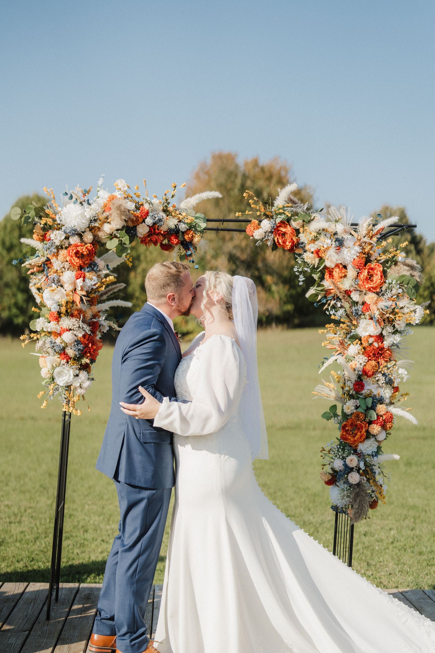 Outdoor wedding ceremony setup with floral arch and couple sharing intimate moment in navy suit and white gown.