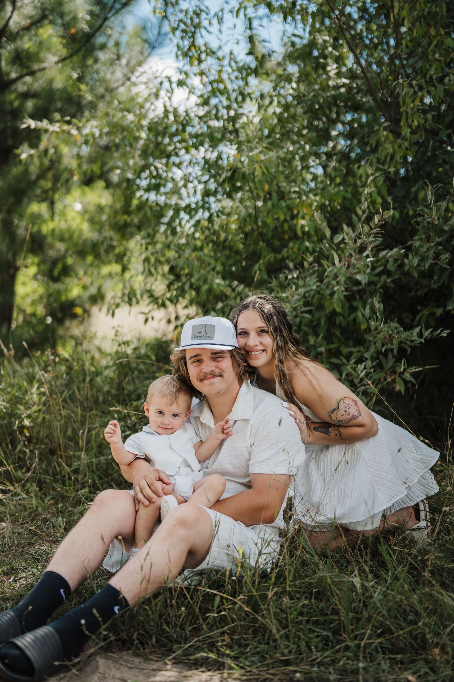 A family in white outfits lounges together on grassy ground surrounded by green foliage on a sunny day.