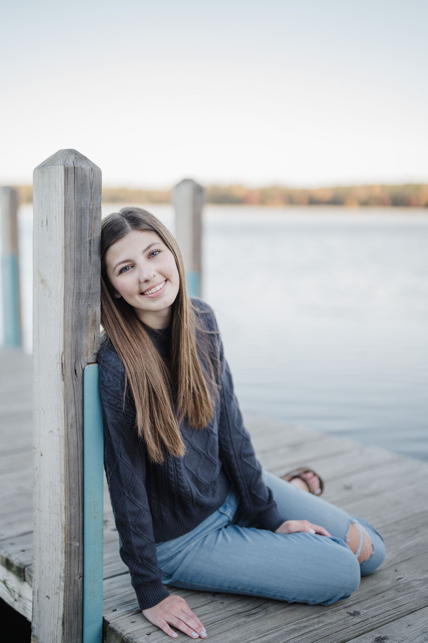 A person in a navy sweater and jeans sits on a wooden dock by calm water at sunset.
