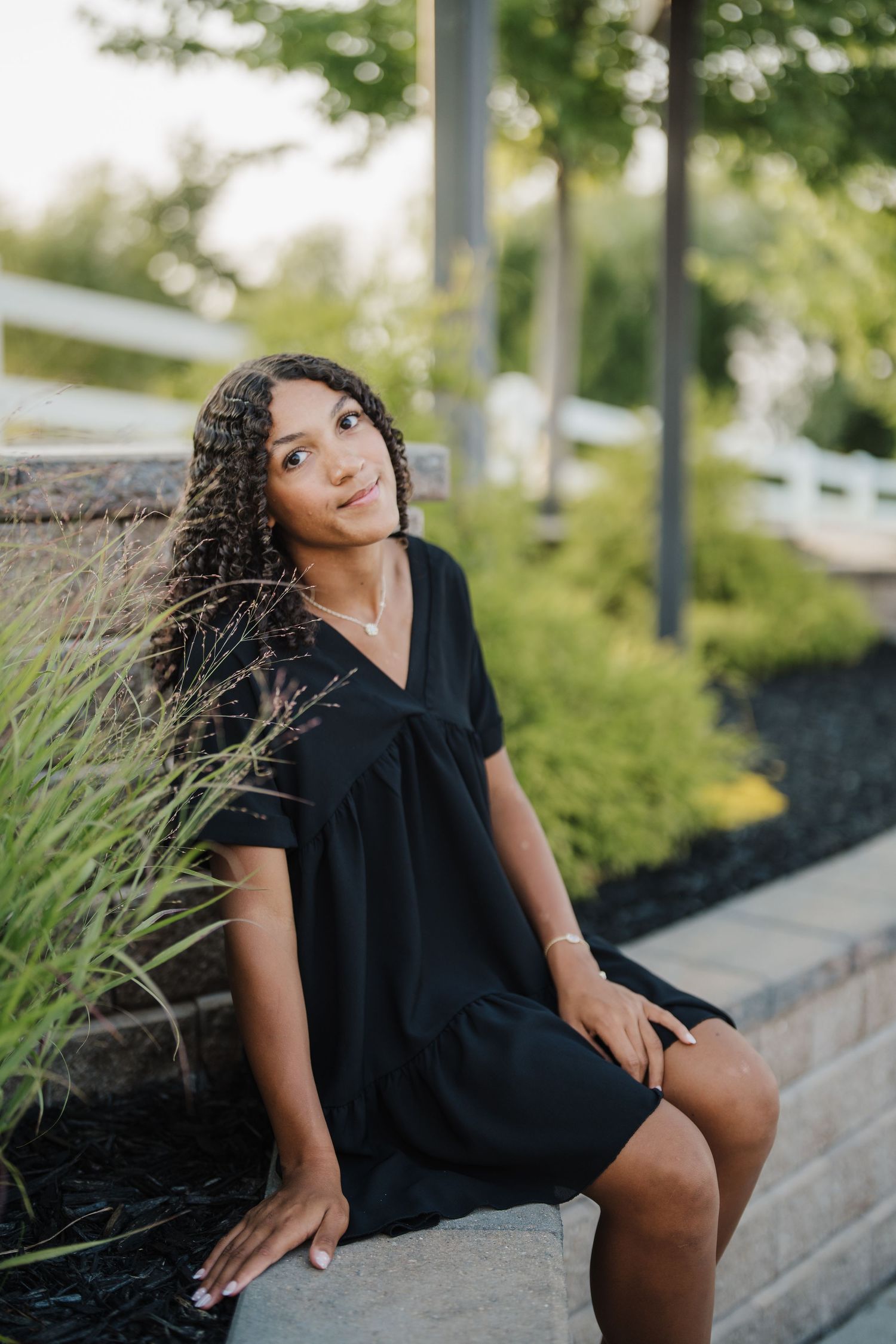A person in a black dress poses outdoors on concrete steps near ornamental grasses in a natural setting.