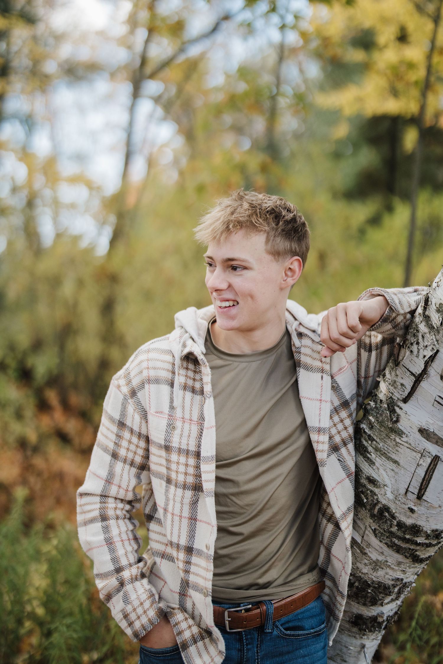 Person in plaid flannel and olive shirt poses casually against birch tree in autumn forest setting.