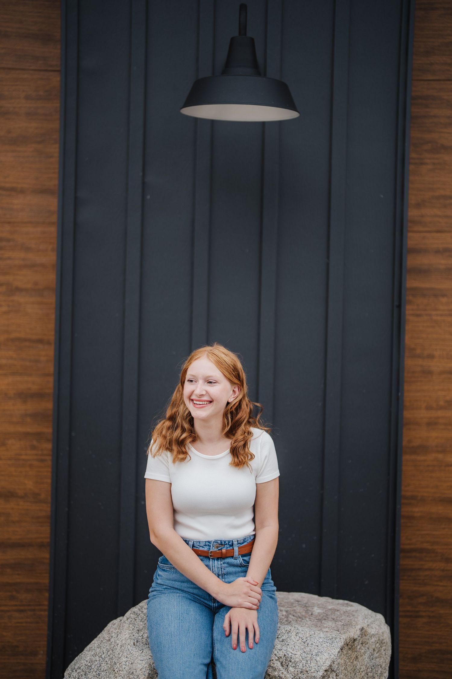 Person in white t-shirt and jeans sitting on rock against dark wall with industrial pendant light above.