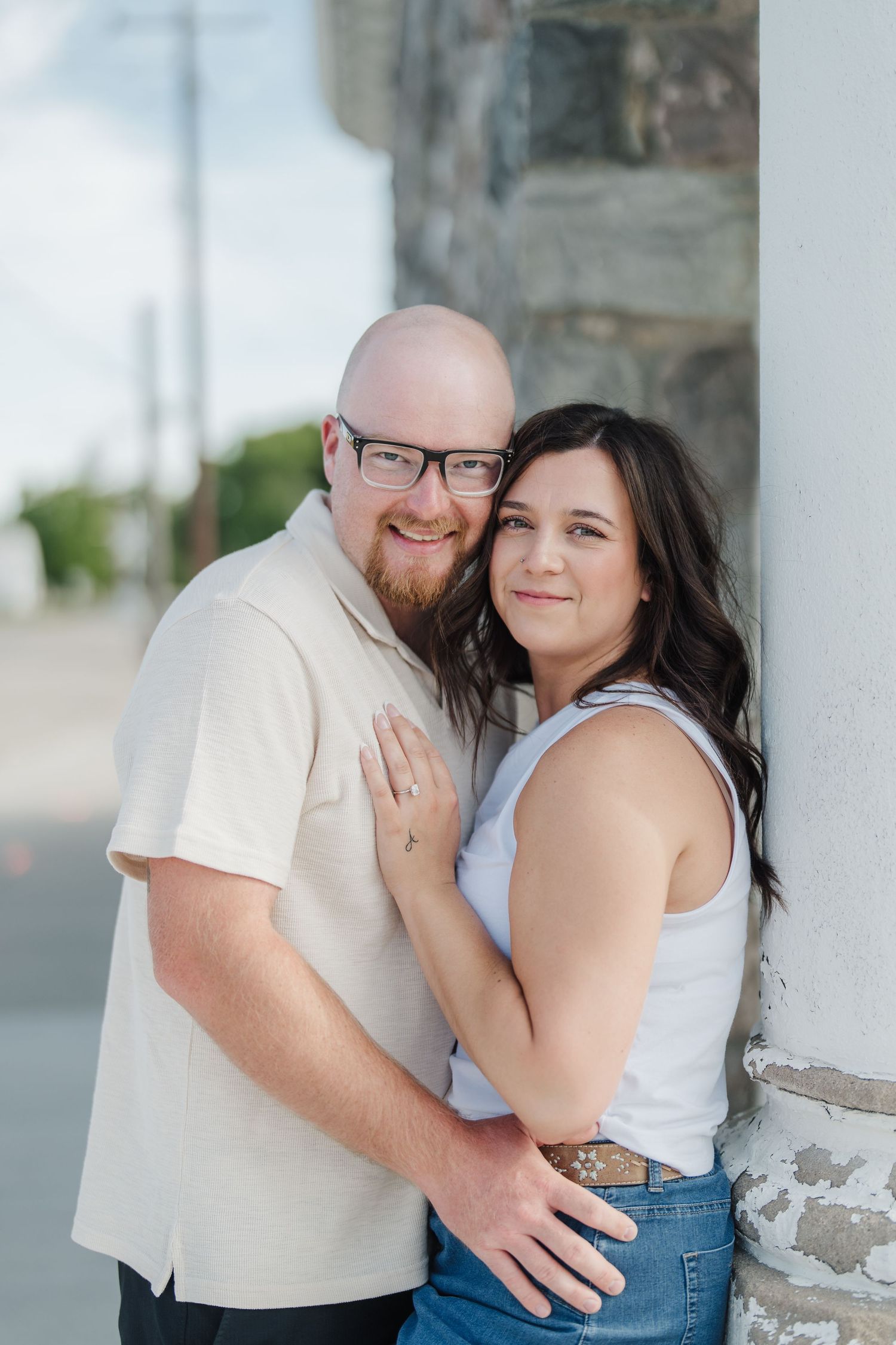 A bald man with glasses and a woman with dark hair embrace and smile outdoors near a stone pillar.