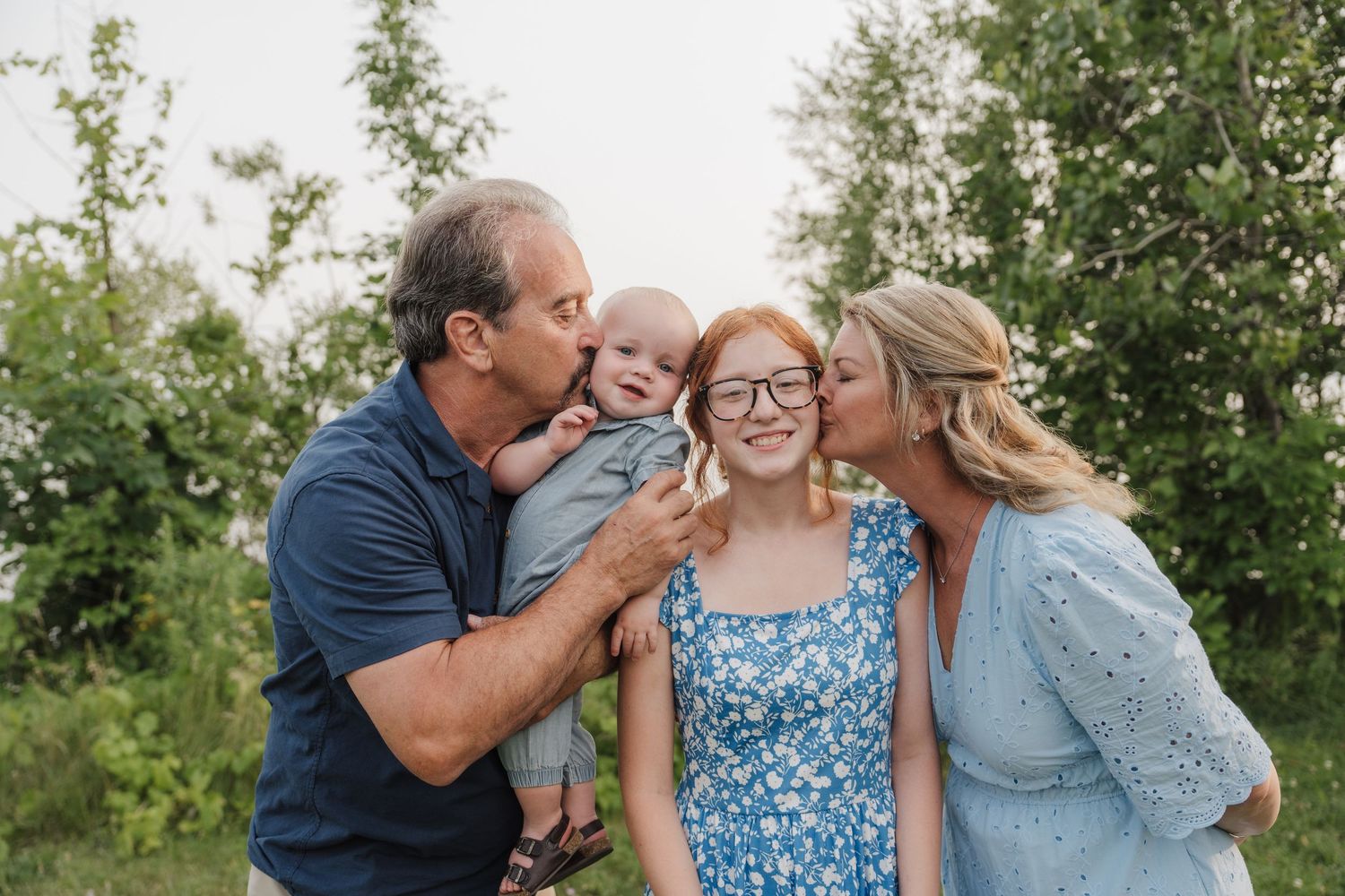 Family of four outdoors, parents kissing teenage girl and baby, surrounded by lush green trees.