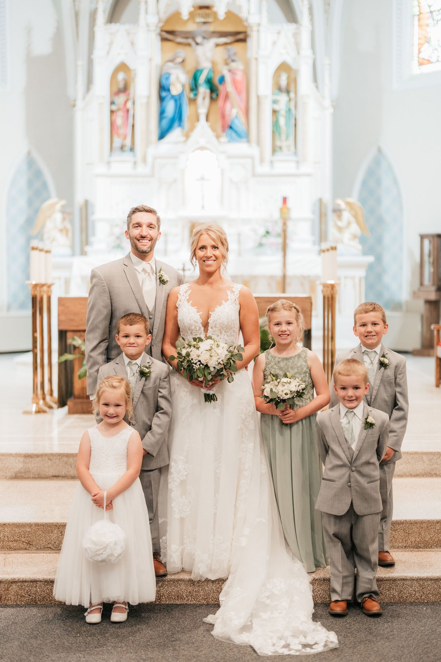 Wedding party poses on church altar steps with flower bouquets in light gray attire.