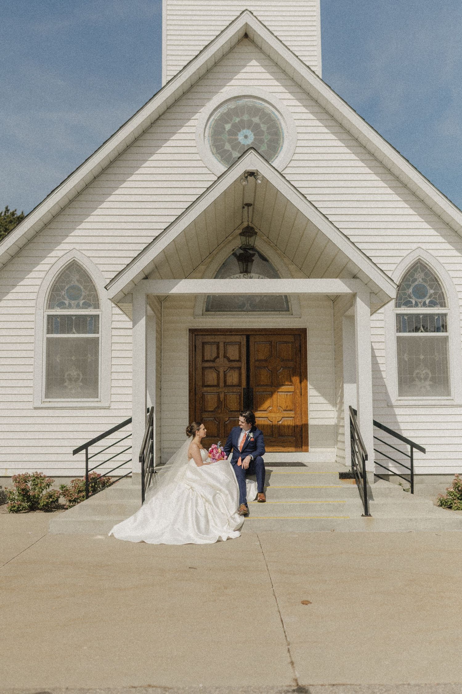 A white church exterior with gothic windows and steps leading to wooden double doors on a sunny day.