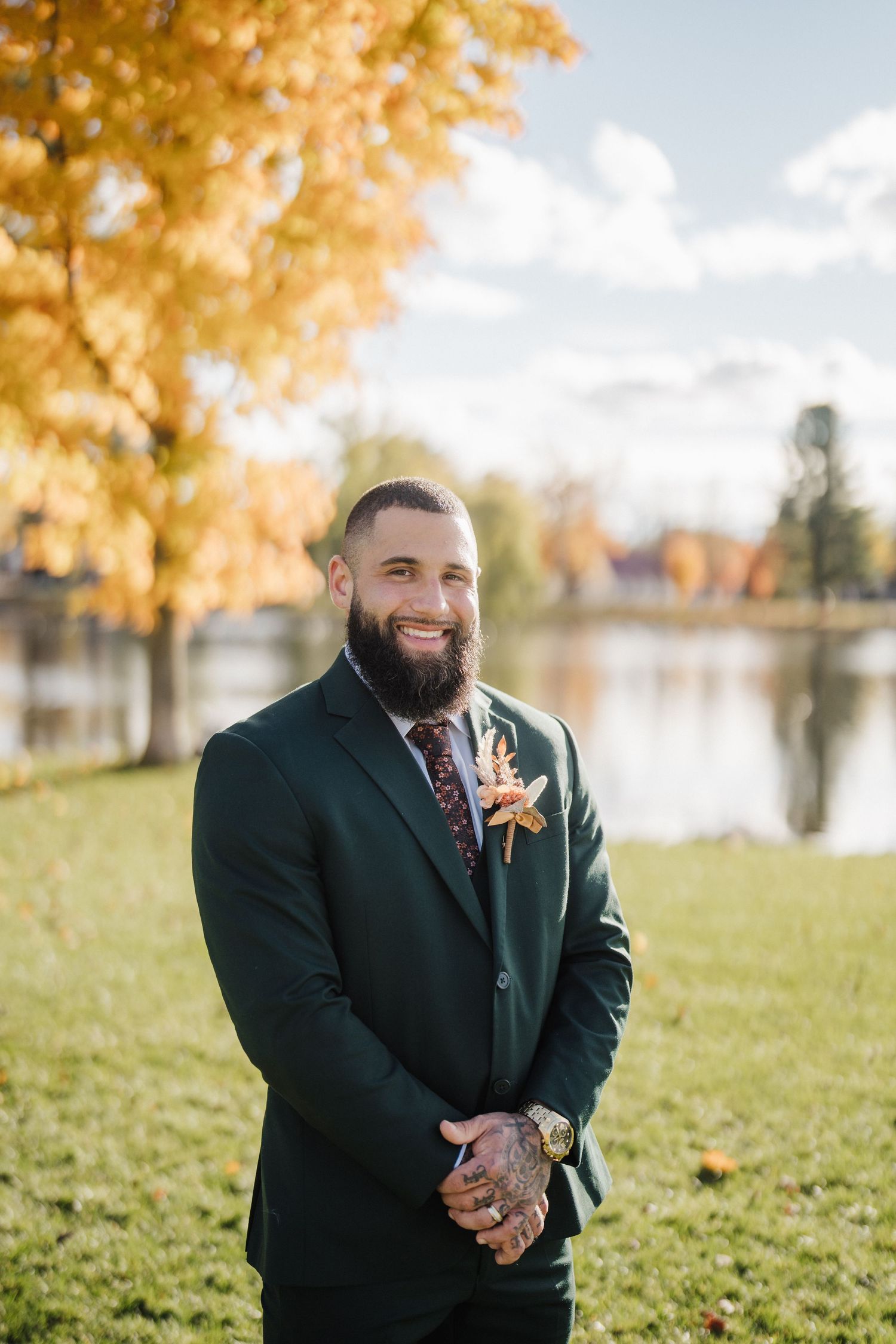 Outdoor autumn wedding portrait with lake and golden trees in background, featuring dark suit and floral boutonniere.