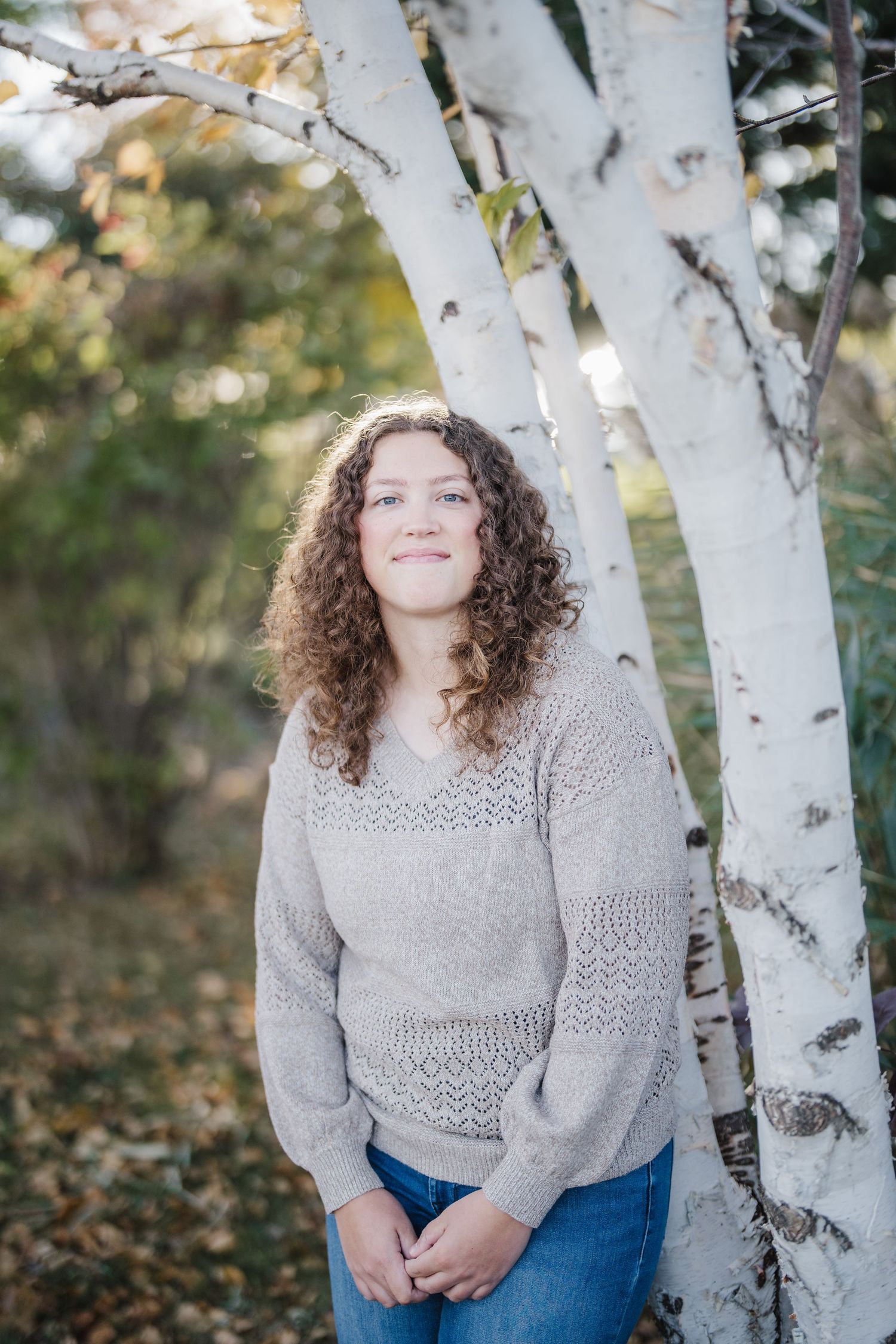 Person in gray sweater and jeans sits near white birch trees during autumn photoshoot.