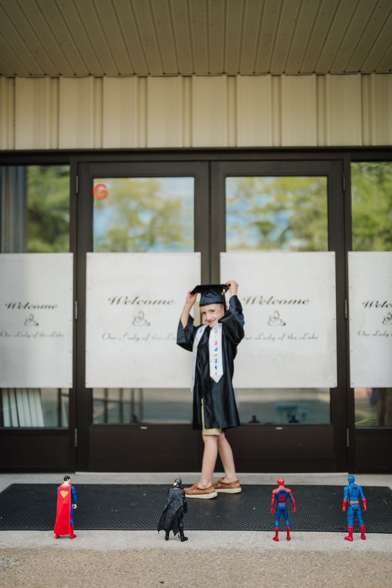 A graduate poses in front of welcome signs with superhero action figures lined up at their feet.