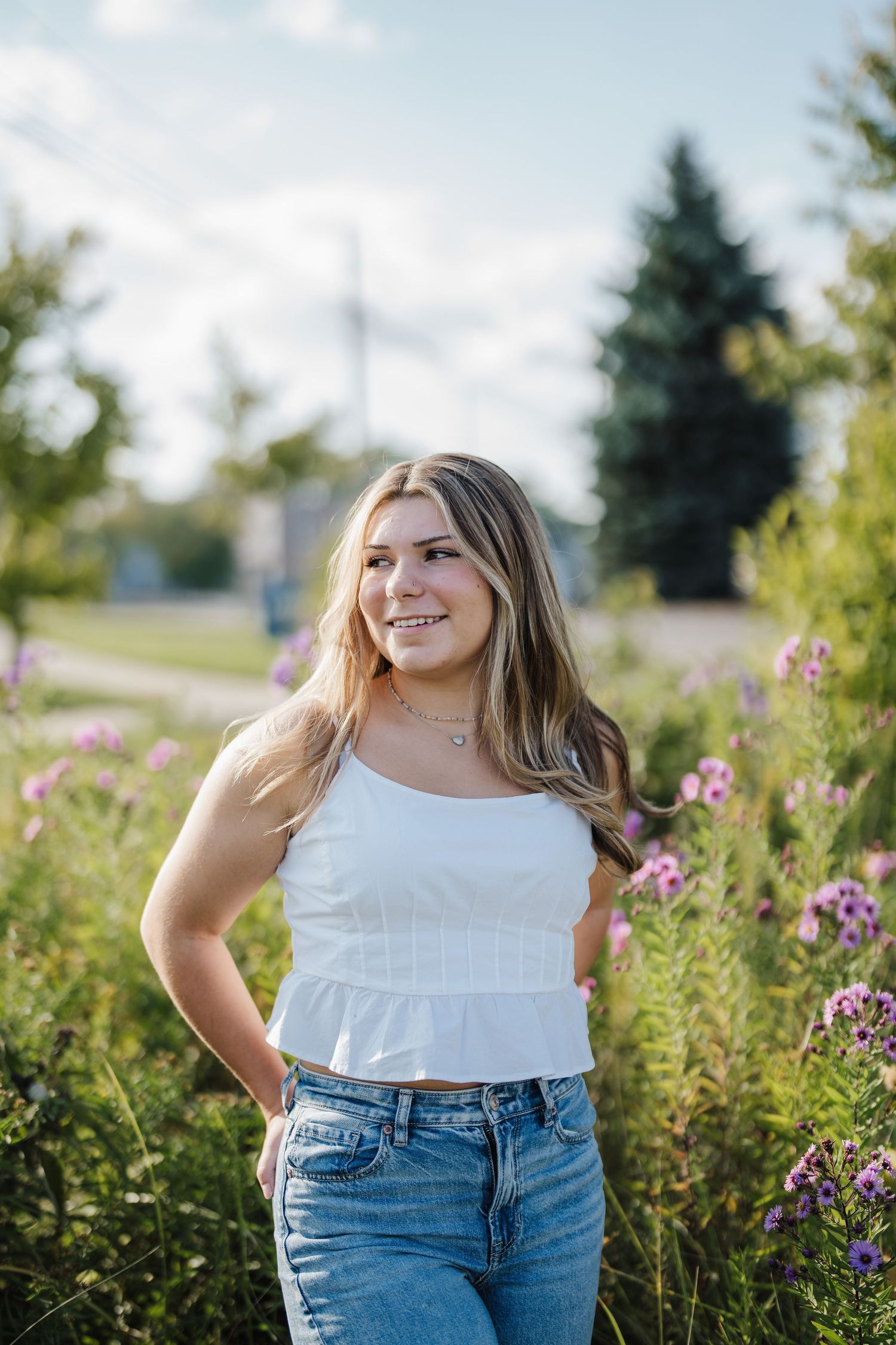 A person in a white top and jeans poses in a field of wildflowers on a sunny summer day.