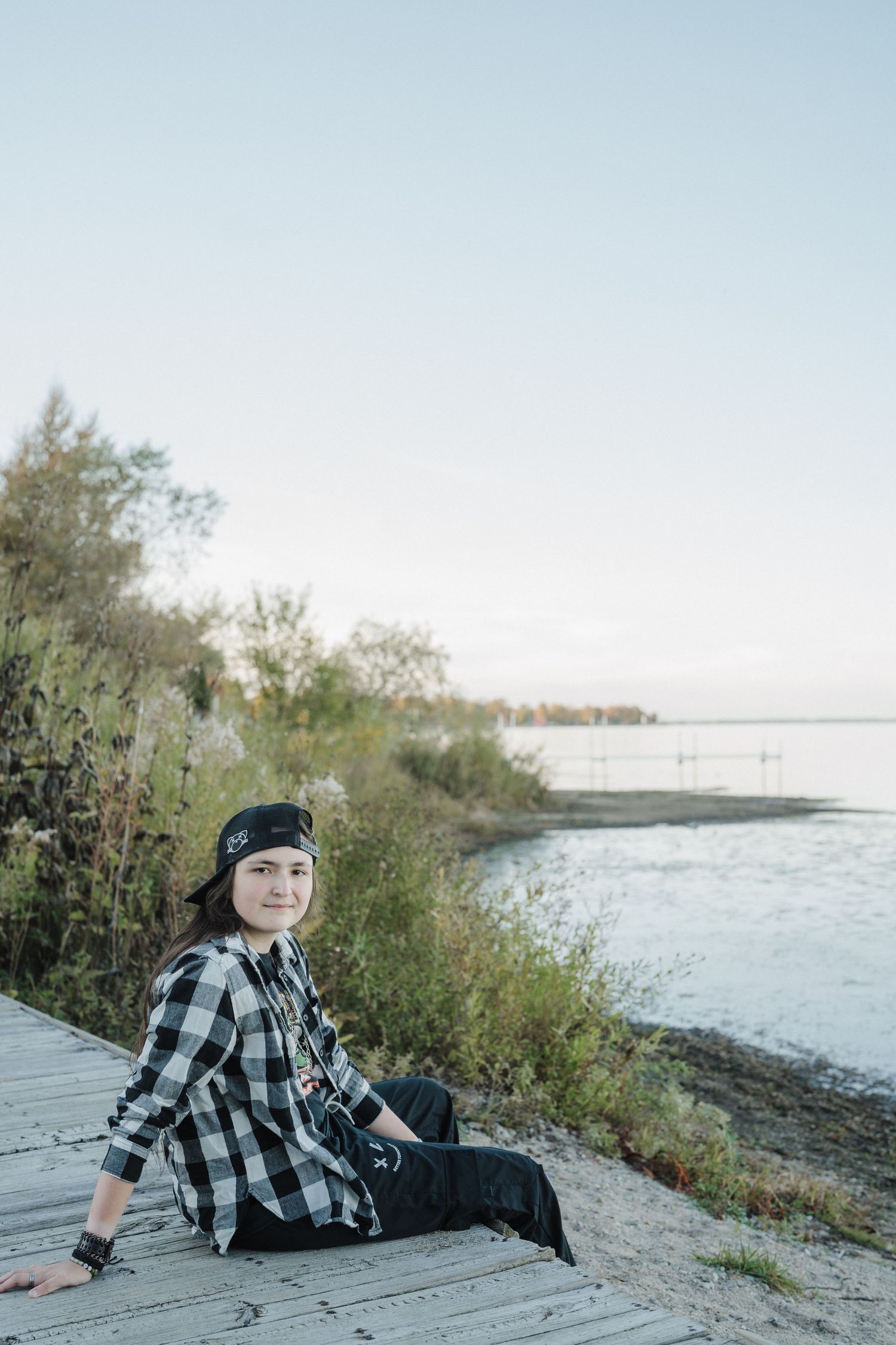 A person in a plaid shirt and beanie sits on a sandy beach near water and trees during golden hour.