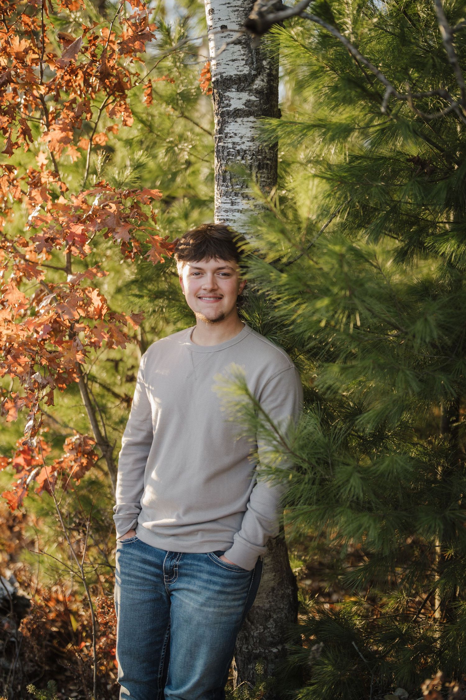Someone in a gray sweater and jeans poses against a backdrop of autumn trees and birch bark.