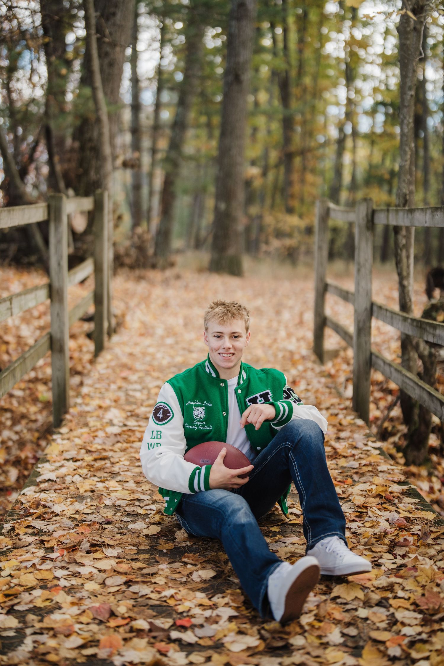 Football player in green and white varsity jacket poses on fallen autumn leaves near wooden fence.