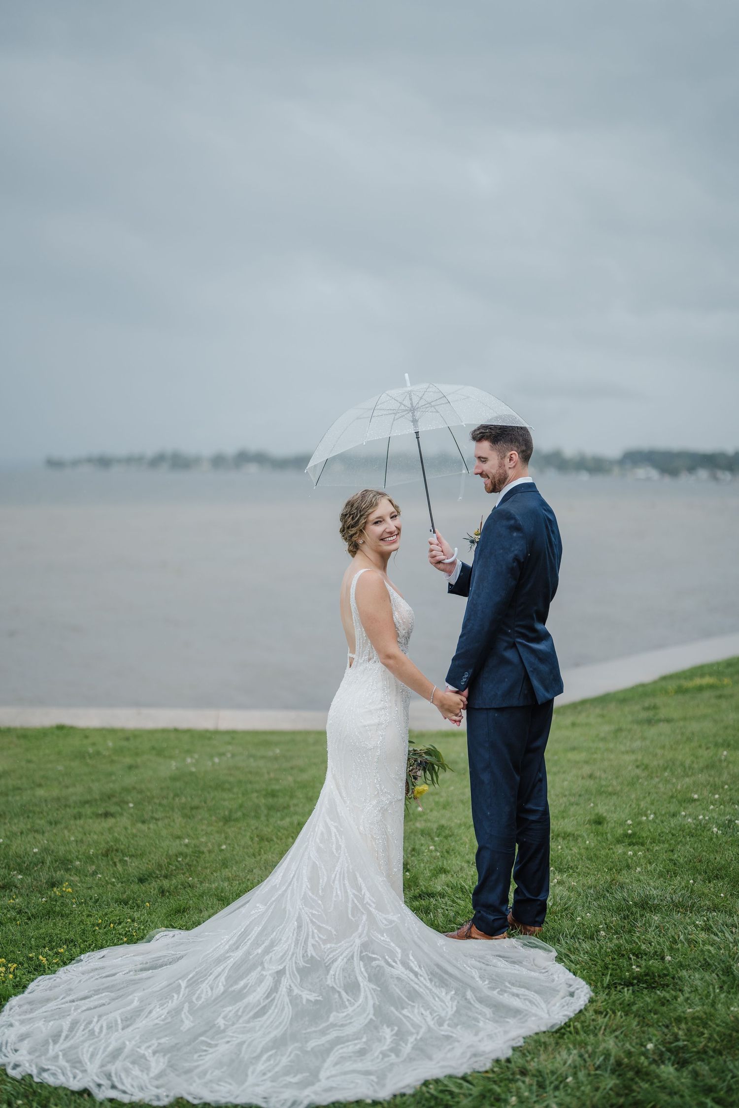 A couple shares an intimate moment under a clear umbrella by a misty waterfront.