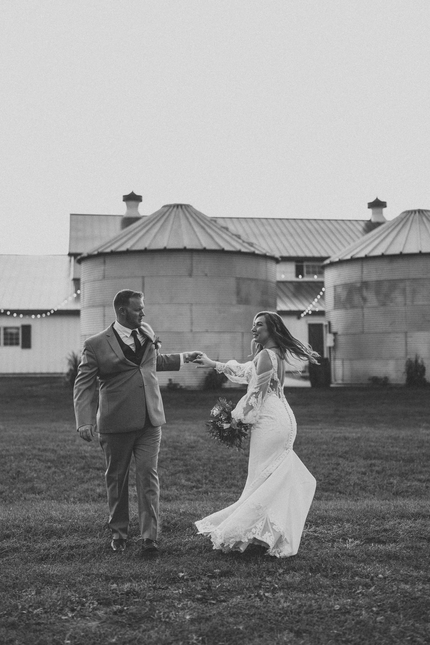 Wedding couple dances playfully in a field with rustic silos in the background.