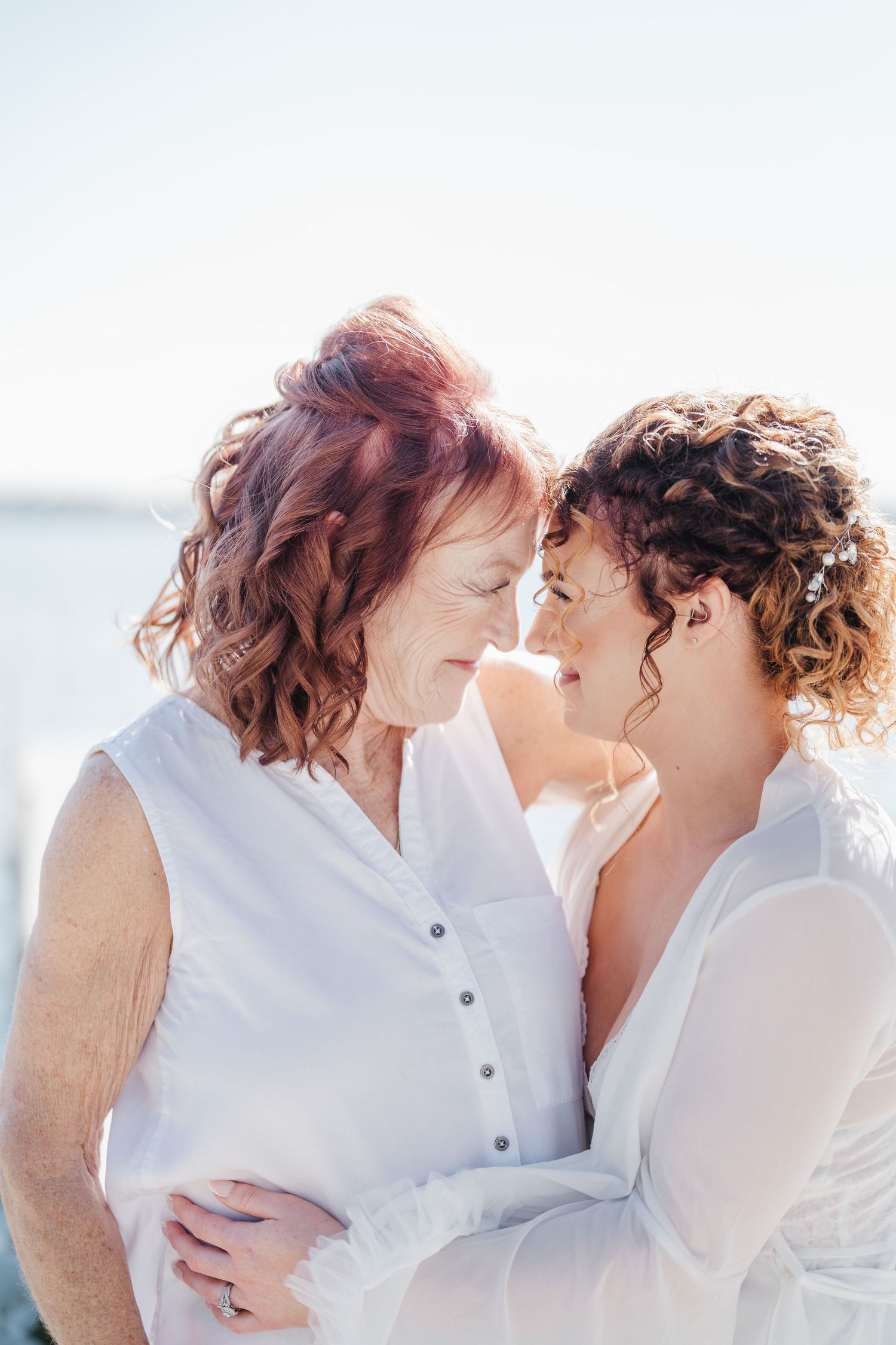 Two people in white outfits share an intimate moment against a waterfront backdrop.