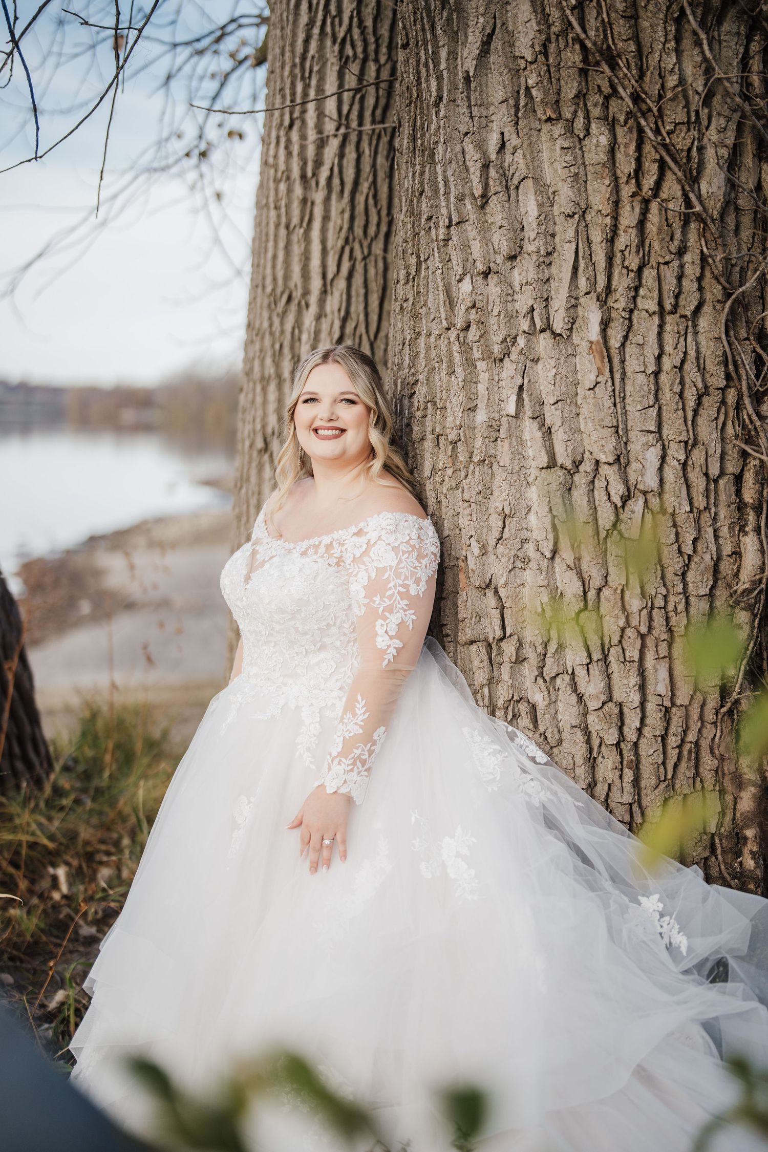 Bride in lace wedding dress poses against large tree trunk by waterfront.