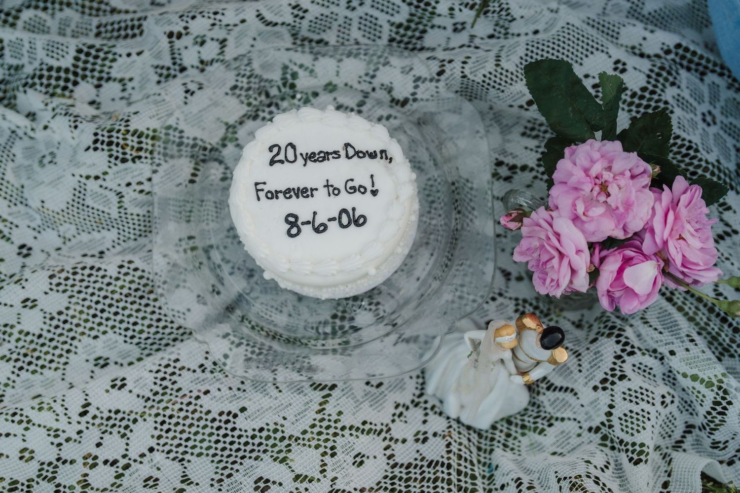 White anniversary cake reading '20 Years Down, Forever to Go! 8-6-06' on lace with pink roses.