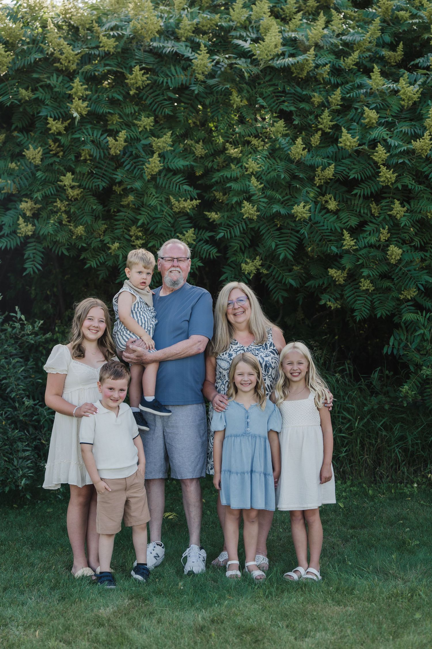 Grandparents posing outdoors with five grandchildren in front of lush green shrubs during a family portrait session.